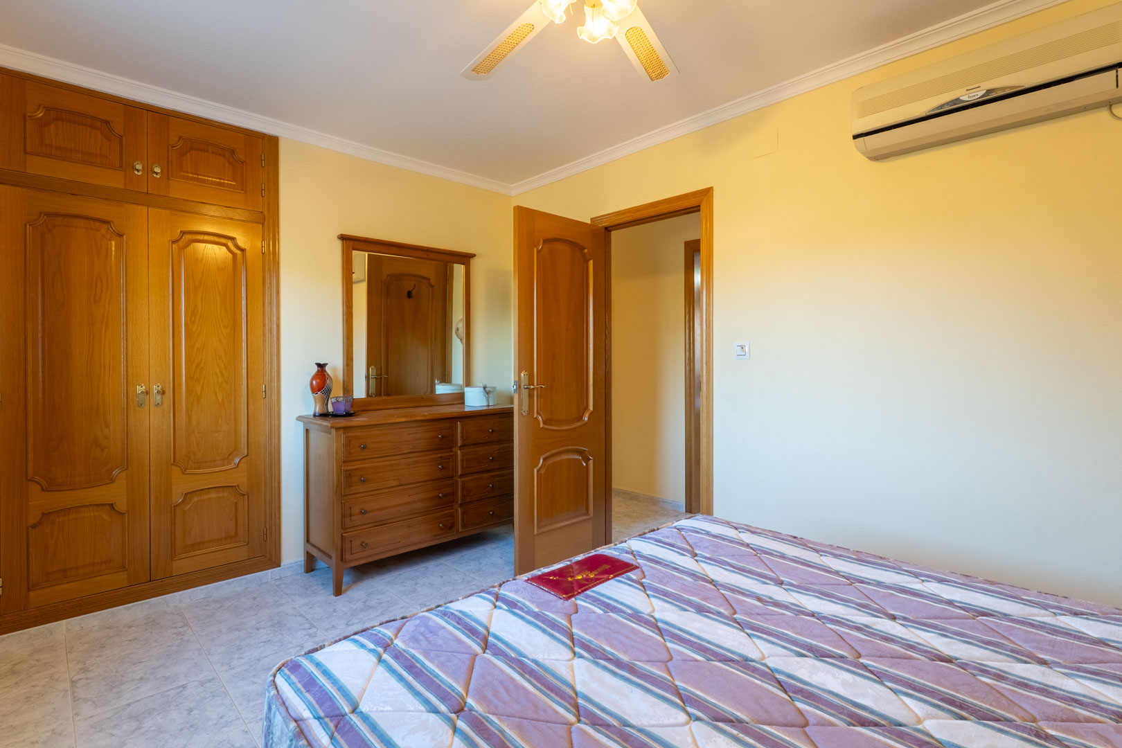 Dormitorio con armarios empotrados de madera, cómoda a juego con espejo, aire acondicionado y ventilador de techo sobre suelos cerámicos claros.