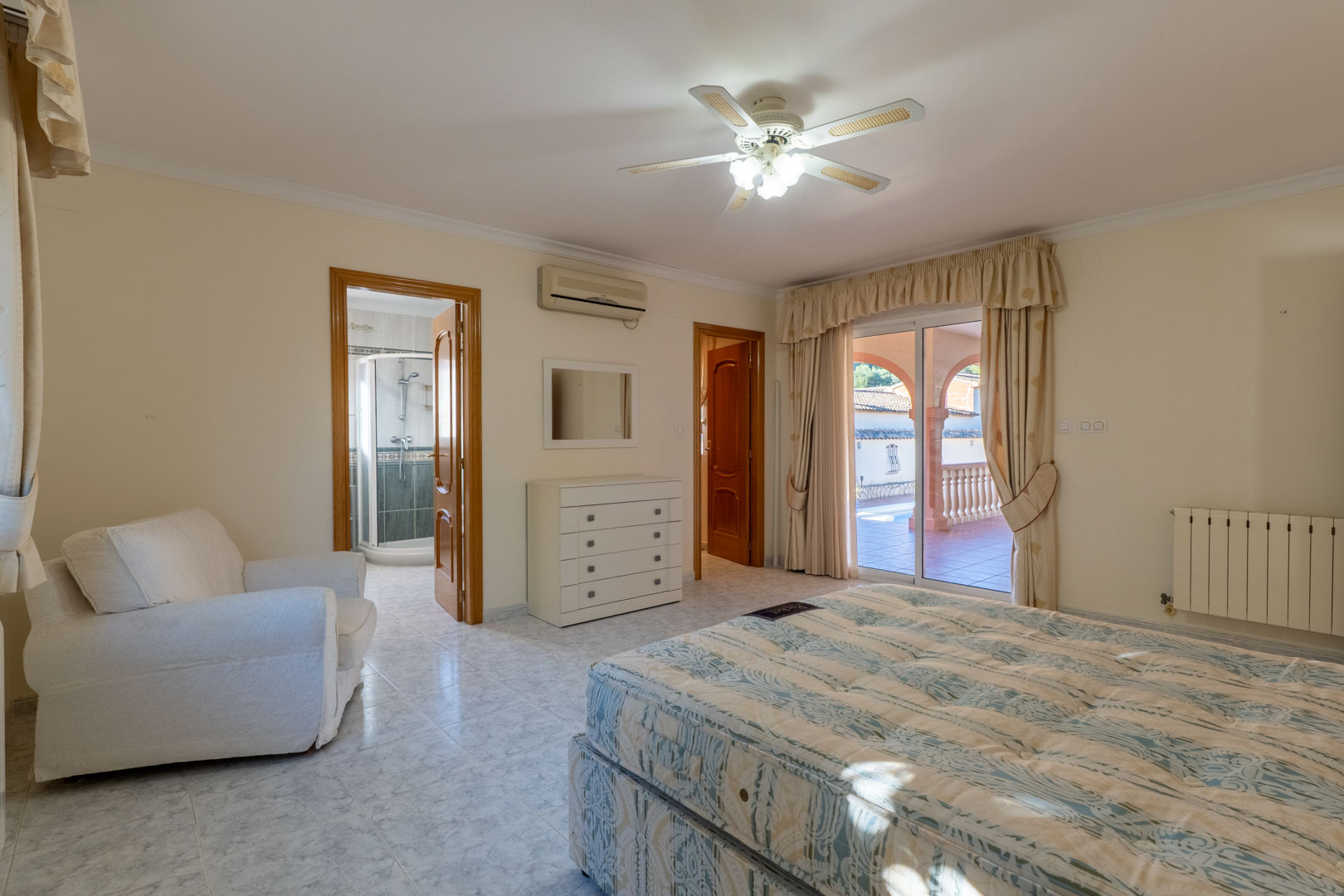Dormitorio principal con baño en suite, aire acondicionado y acceso directo a la terraza. Incluye suelos cerámicos claros, ventilador de techo y vistas al área de la piscina.