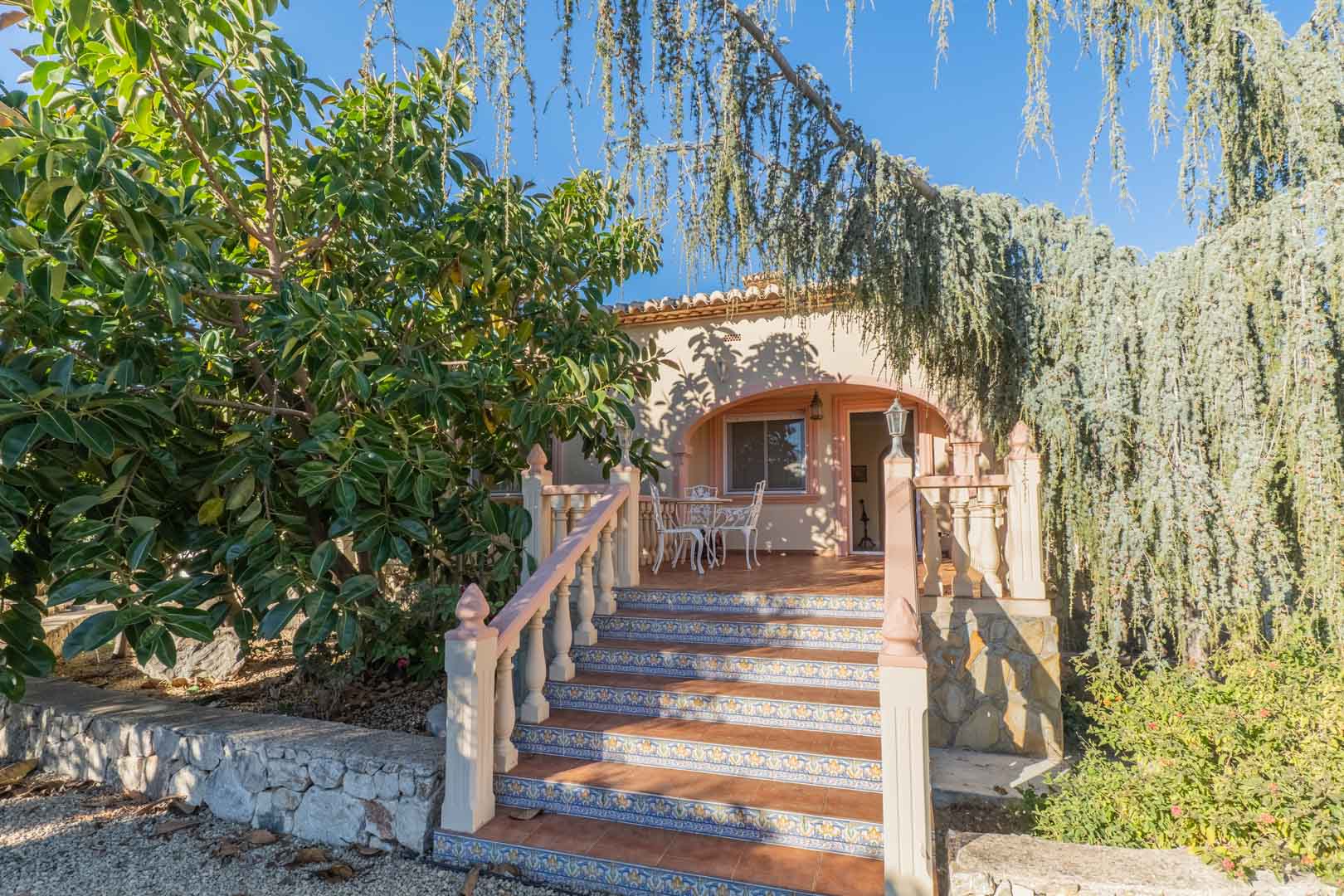 Escalera exterior con azulejos decorativos que conduce a una naya arqueada, rodeada de vegetación madura y un área de descanso con mobiliario de forja blanca.
