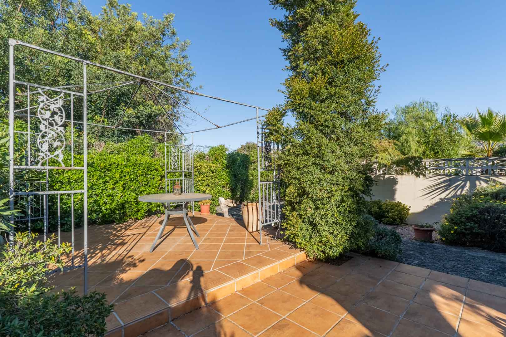 Rincón exterior con suelo de baldosas de barro, pérgola metálica decorativa y mesa auxiliar, rodeado de vegetación mediterránea madura y setos cuidados.