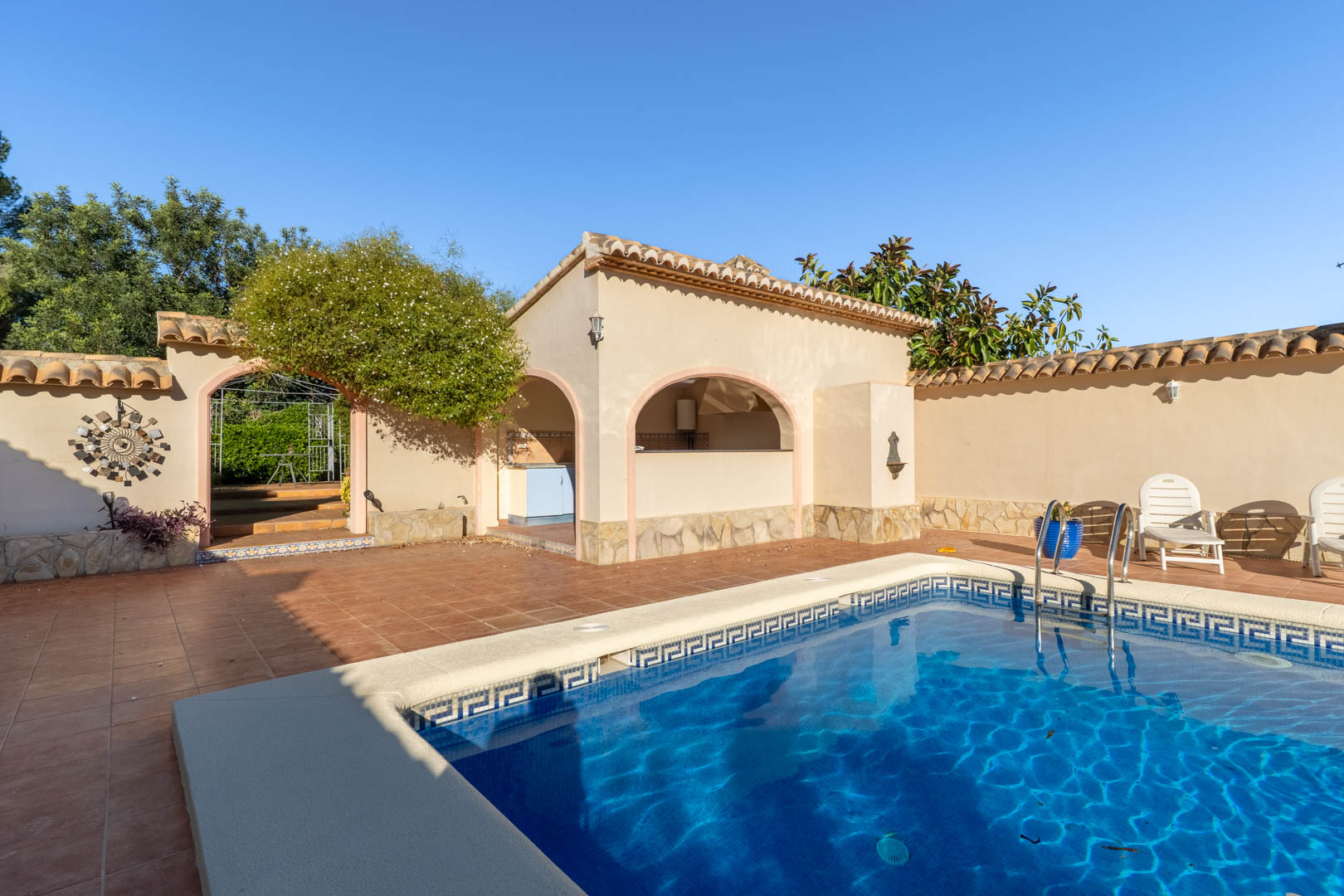 Zona de piscina con terraza de terracota, cocina de verano bajo arcos mediterráneos y acceso decorativo al jardín exterior.
