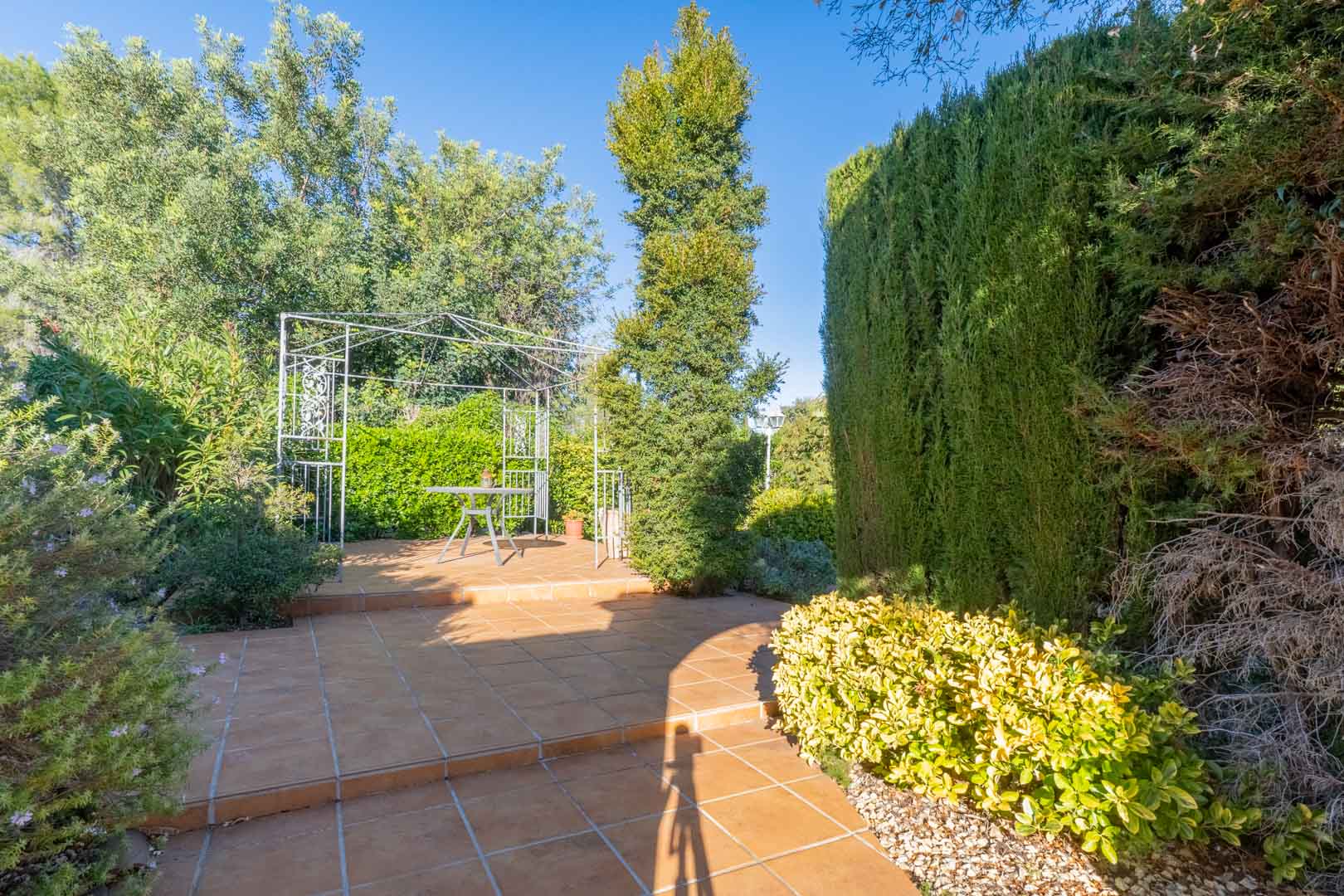 Zona de terraza exterior con suelo de gres cerámico, pérgola metálica blanca y vegetación mediterránea madura que proporciona privacidad natural.