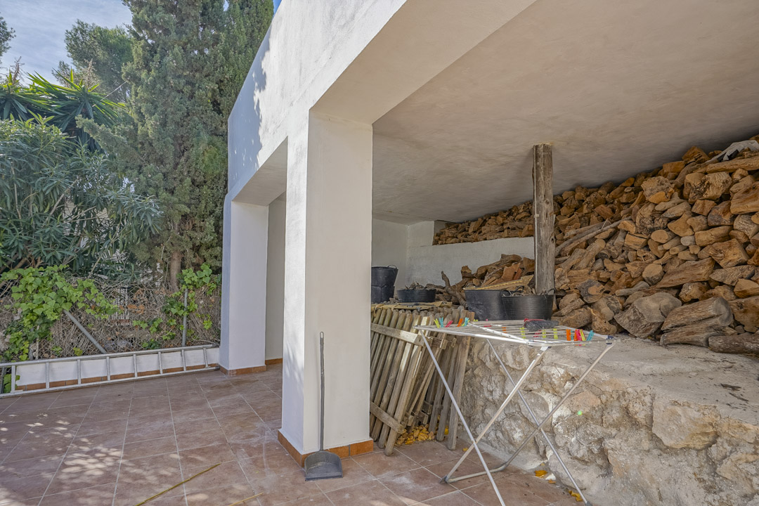 Área de almacenamiento exterior cubierta con suelo de baldosas cerámicas, pilares blancos y un gran leñero sobre base de piedra natural.