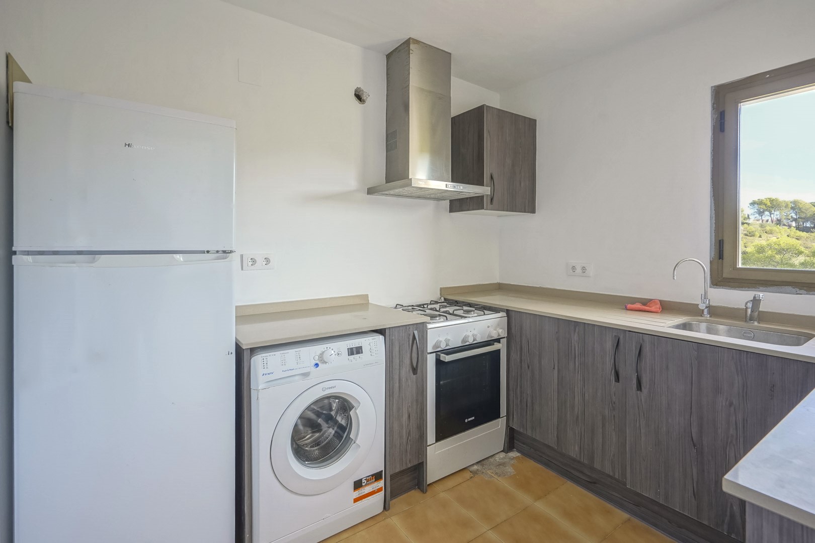 Cocina funcional con armarios en acabado madera gris, equipada con electrodomésticos, encimera beige y ventana con vistas despejadas al entorno natural.