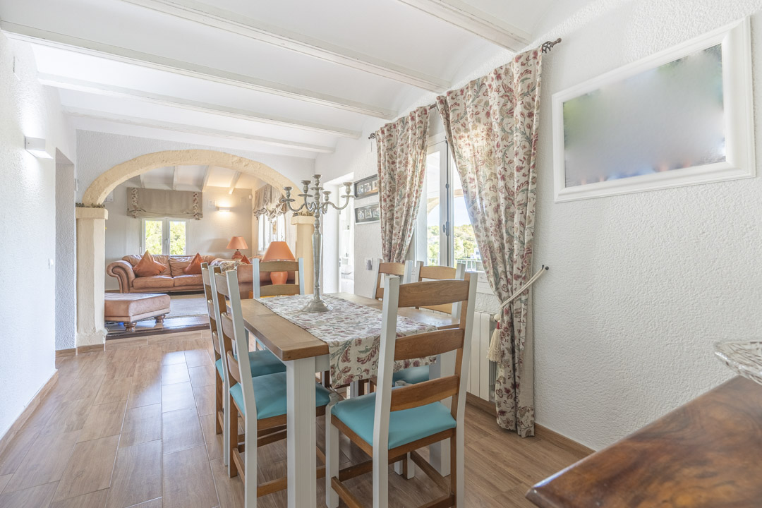 Comedor luminoso con vigas blancas y arco de piedra tosca que comunica con el salón. Suelos cerámicos de efecto madera y mobiliario de estilo rústico.