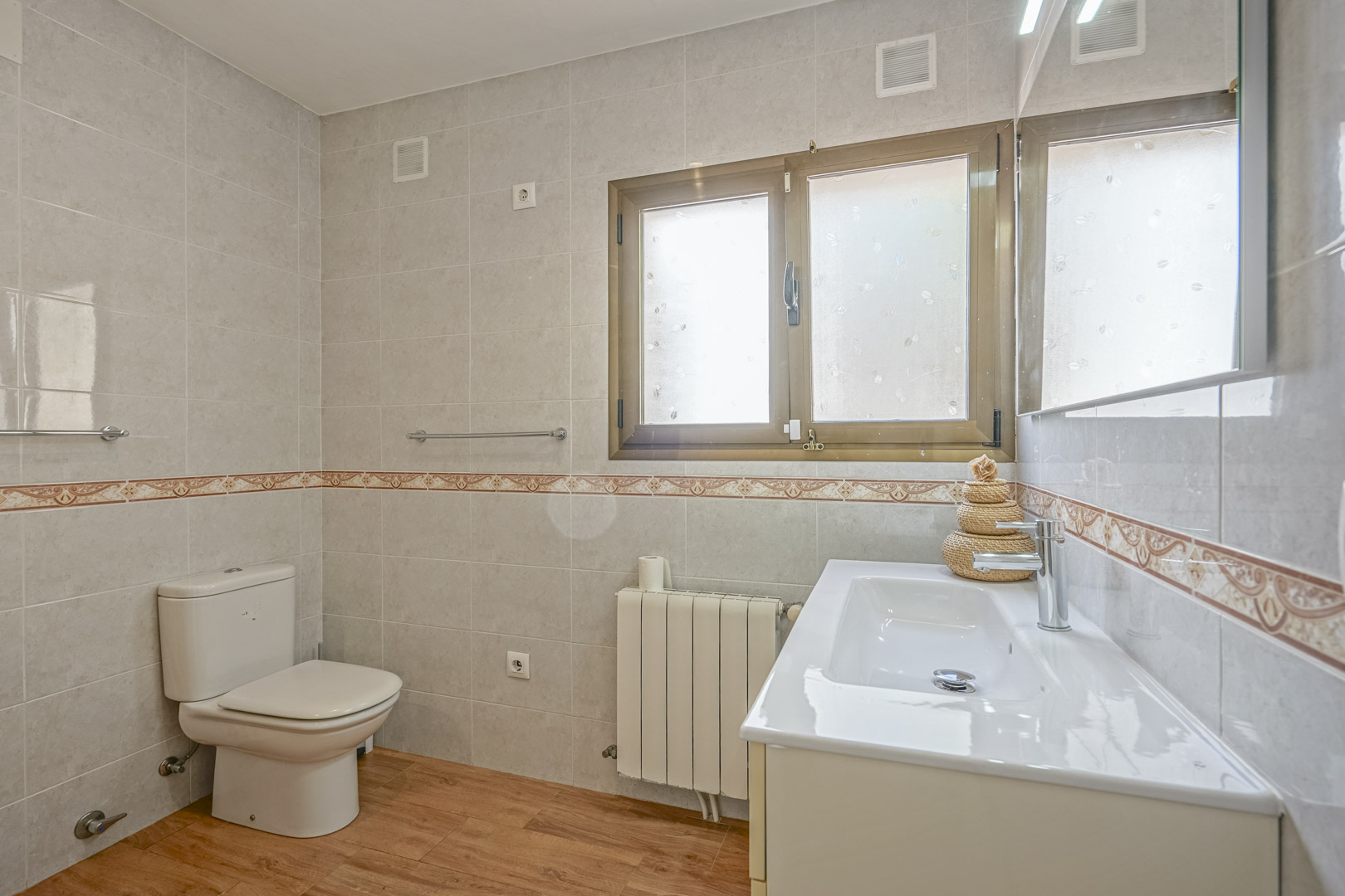 Cuarto de baño reformado con suelos de efecto madera, paredes alicatadas con cenefa decorativa, radiador y mueble de lavabo blanco con espejo.