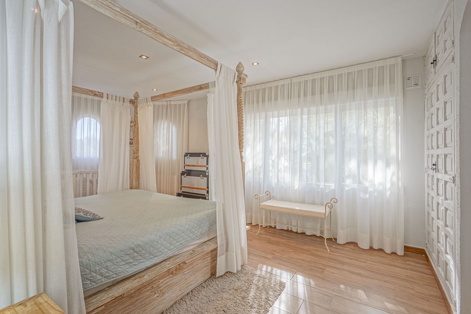Dormitorio con cama de dosel en madera clara, suelos cerámicos de efecto madera y amplios ventanales con cortinas traslúcidas que filtran la luz natural.