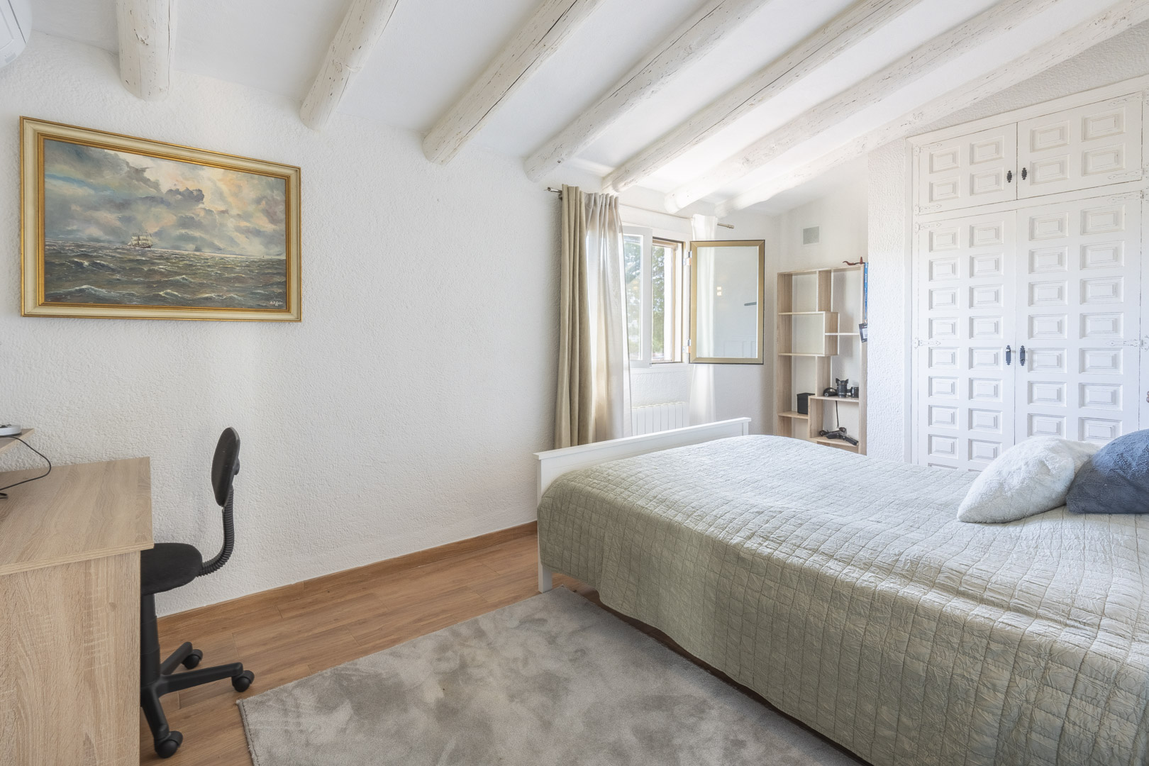 Dormitorio con vigas de madera blancas, techos inclinados, armarios empotrados de estilo tradicional y zona de escritorio con luz natural.