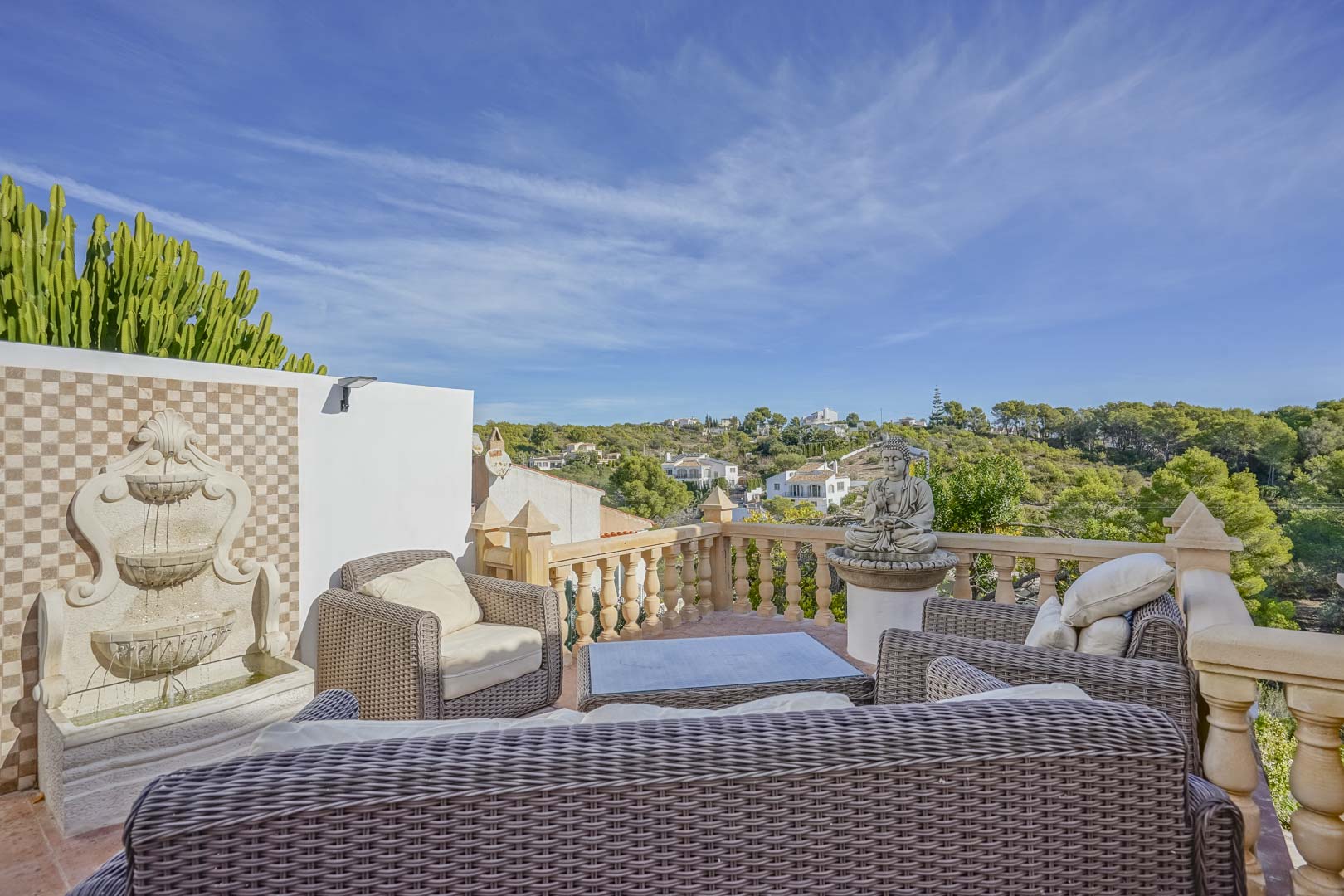 Terraza elevada con mobiliario de mimbre, fuente de pared decorativa y balaustrada de piedra con vistas despejadas al entorno natural de Jávea.