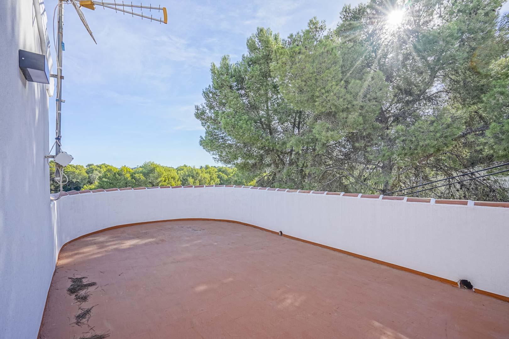 Terraza superior con suelo de baldosas de terracota y muro perimetral curvo, ofreciendo vistas despejadas a la zona boscosa de pinos bajo el sol mediterráneo.