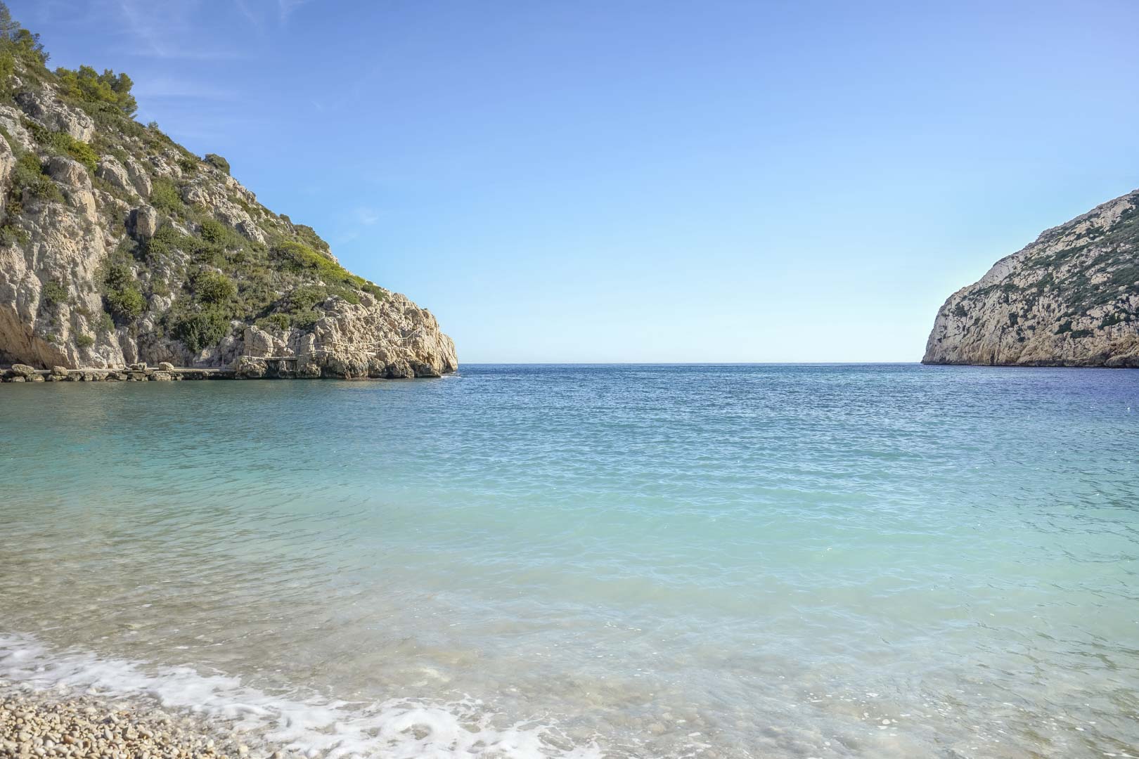Vista de una cala mediterránea de aguas cristalinas y orilla de guijarros, rodeada de acantilados rocosos y situada a pocos minutos de la propiedad en Jávea.