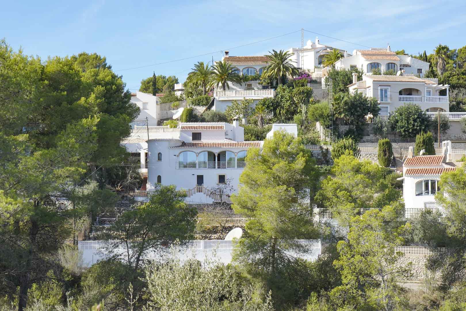Vista exterior de la villa de estilo mediterráneo situada en una ladera arbolada, destacando su fachada blanca, naya acristalada y torreón tradicional.