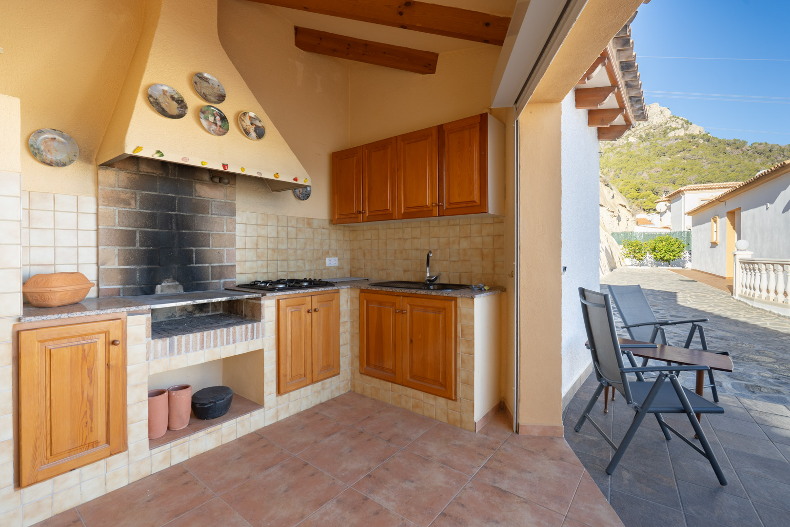 Cocina de verano cubierta con barbacoa de obra, mobiliario de madera, encimera de granito y fregadero, situada junto a una terraza con vistas a la Sierra de Oltà.