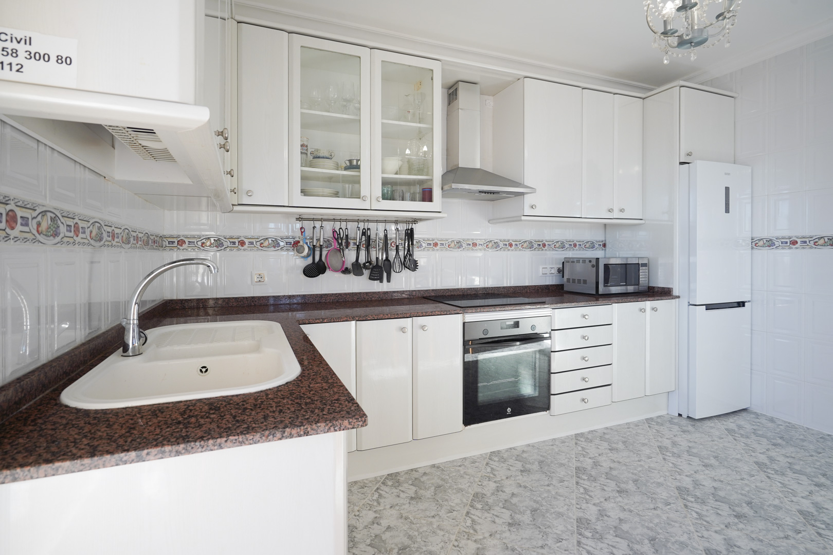 Cocina equipada con armarios blancos, encimera de granito rojizo y azulejos con cenefa decorativa. Incluye electrodomésticos integrados y suelos cerámicos de efecto mármol.