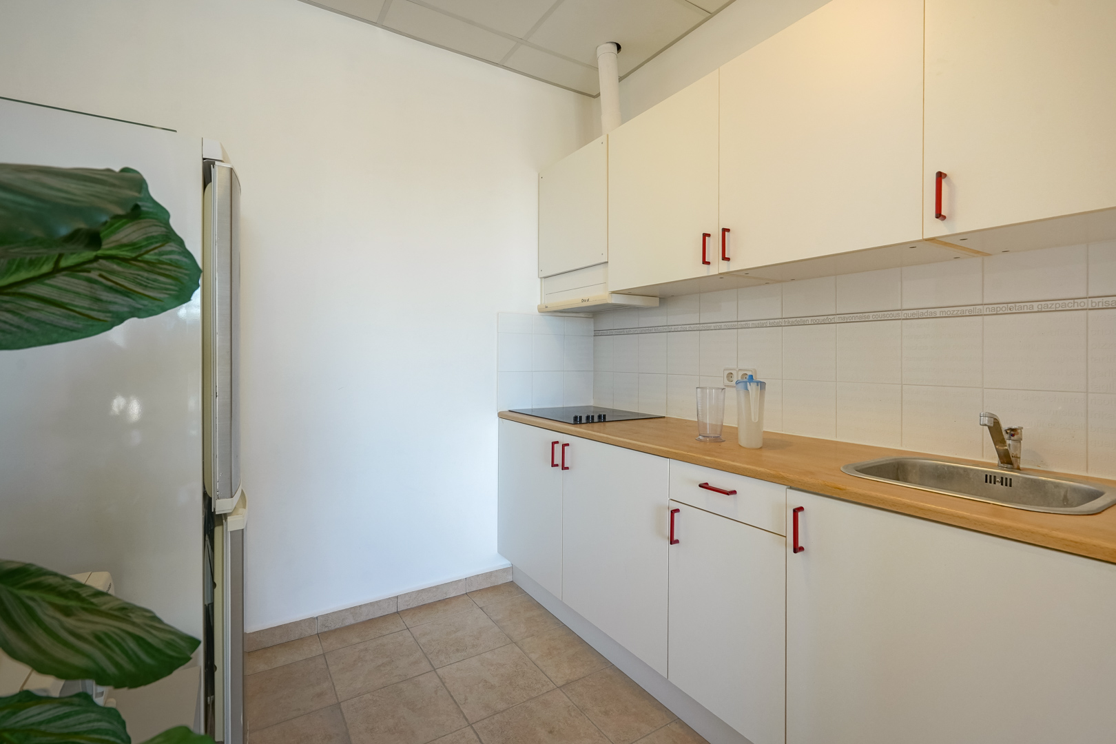 Cocina funcional con armarios blancos, tiradores rojos, encimera de madera y salpicadero de azulejos. Incluye placa de cocción y fregadero de acero inoxidable.
