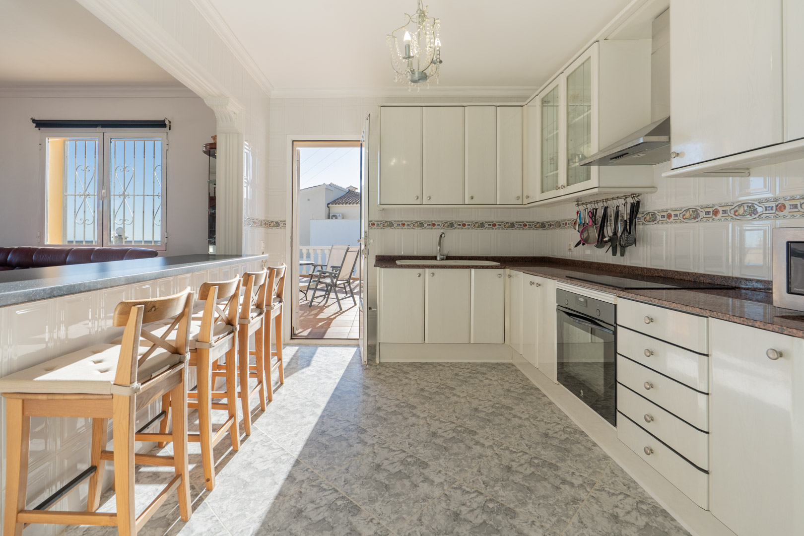 Cocina semiabierta con armarios blancos, encimera de granito y barra con taburetes. Ofrece acceso directo a la terraza y suelos de gres con efecto mármol.