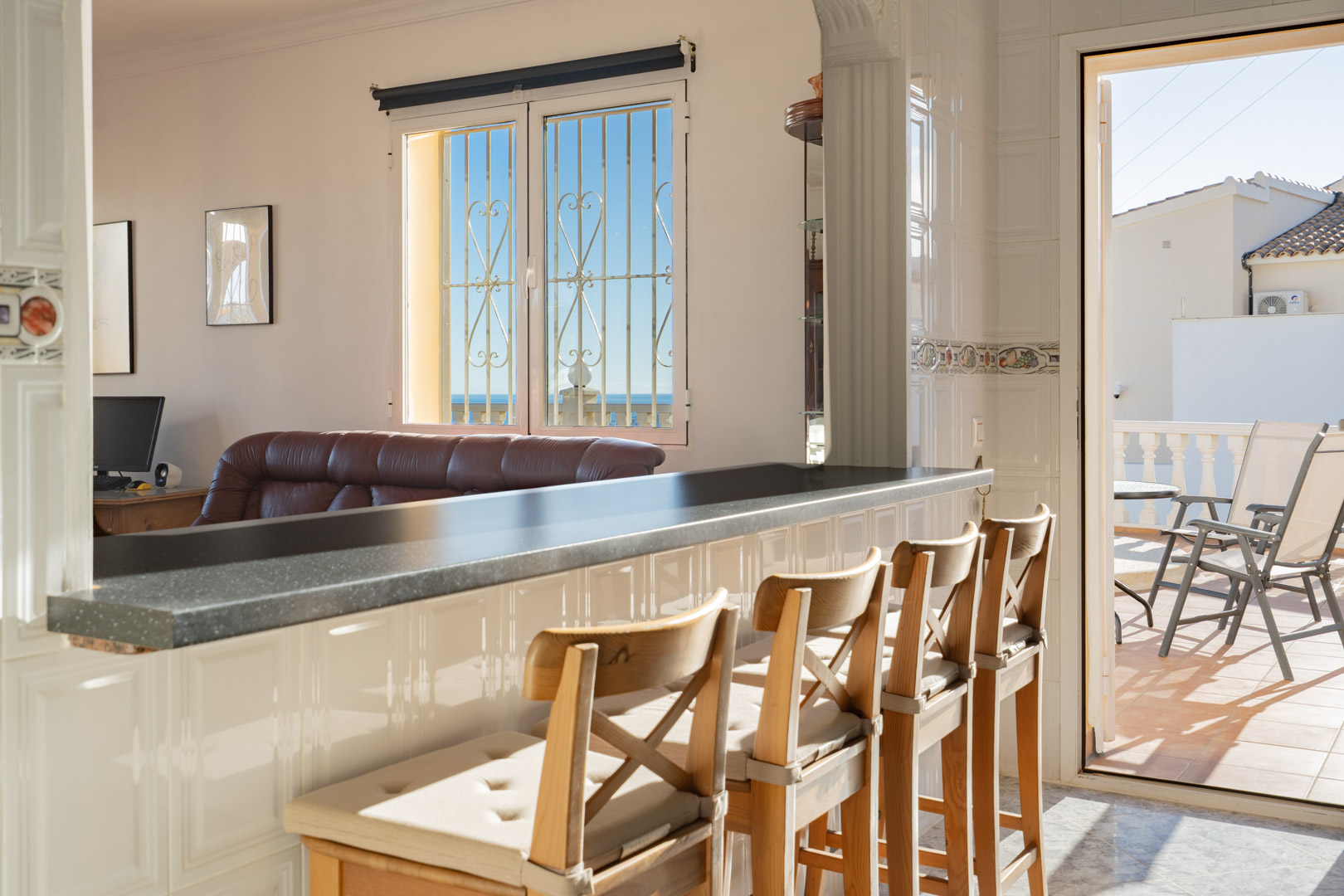 Cocina semiabierta con barra americana de encimera oscura y taburetes de madera, conectada al salón y a una terraza soleada con vistas al mar.