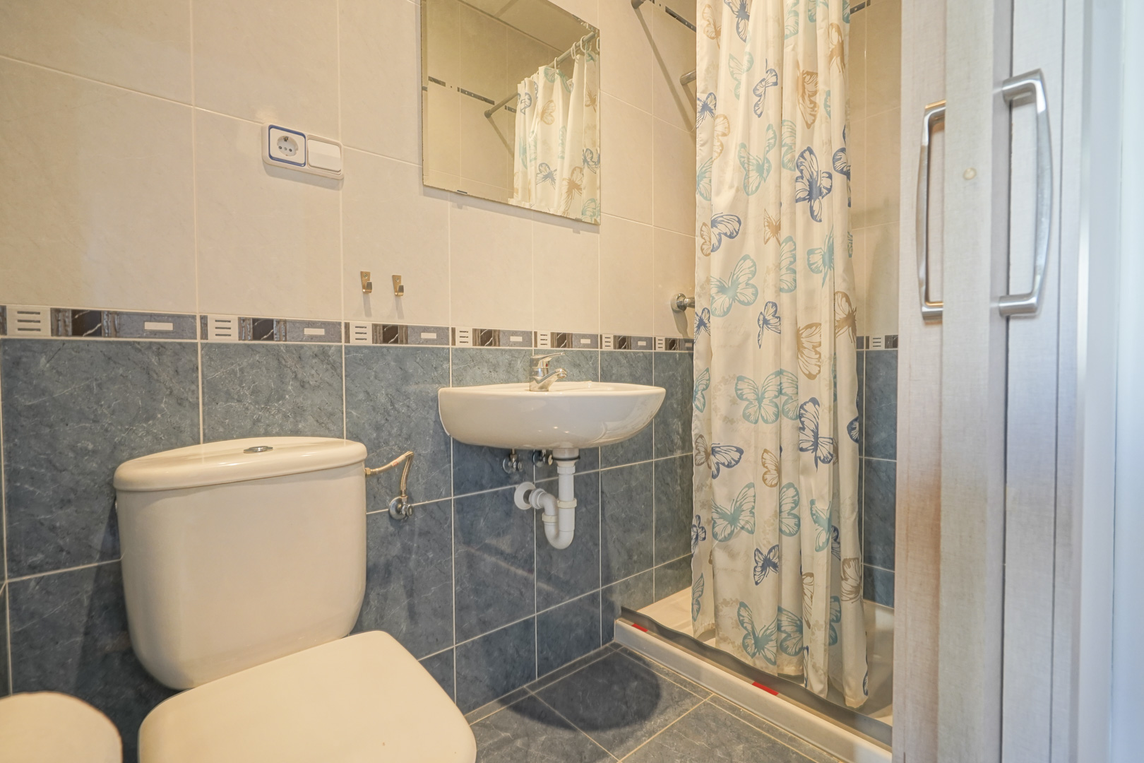 Cuarto de baño con azulejos en tonos azules y crema, equipado con inodoro, lavabo suspendido y zona de ducha con cortina decorativa de mariposas.