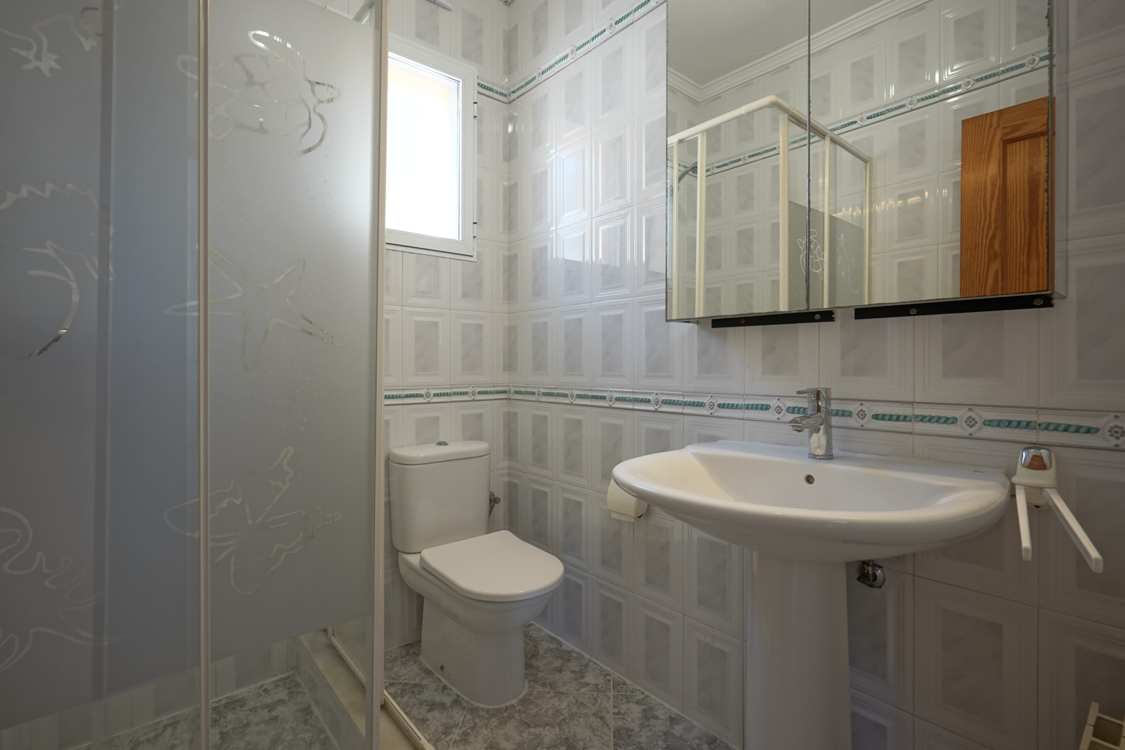 Cuarto de baño con lavabo de pedestal, inodoro y ducha con mampara de cristal decorada. Paredes alicatadas con cenefa ornamental y ventana para luz natural.