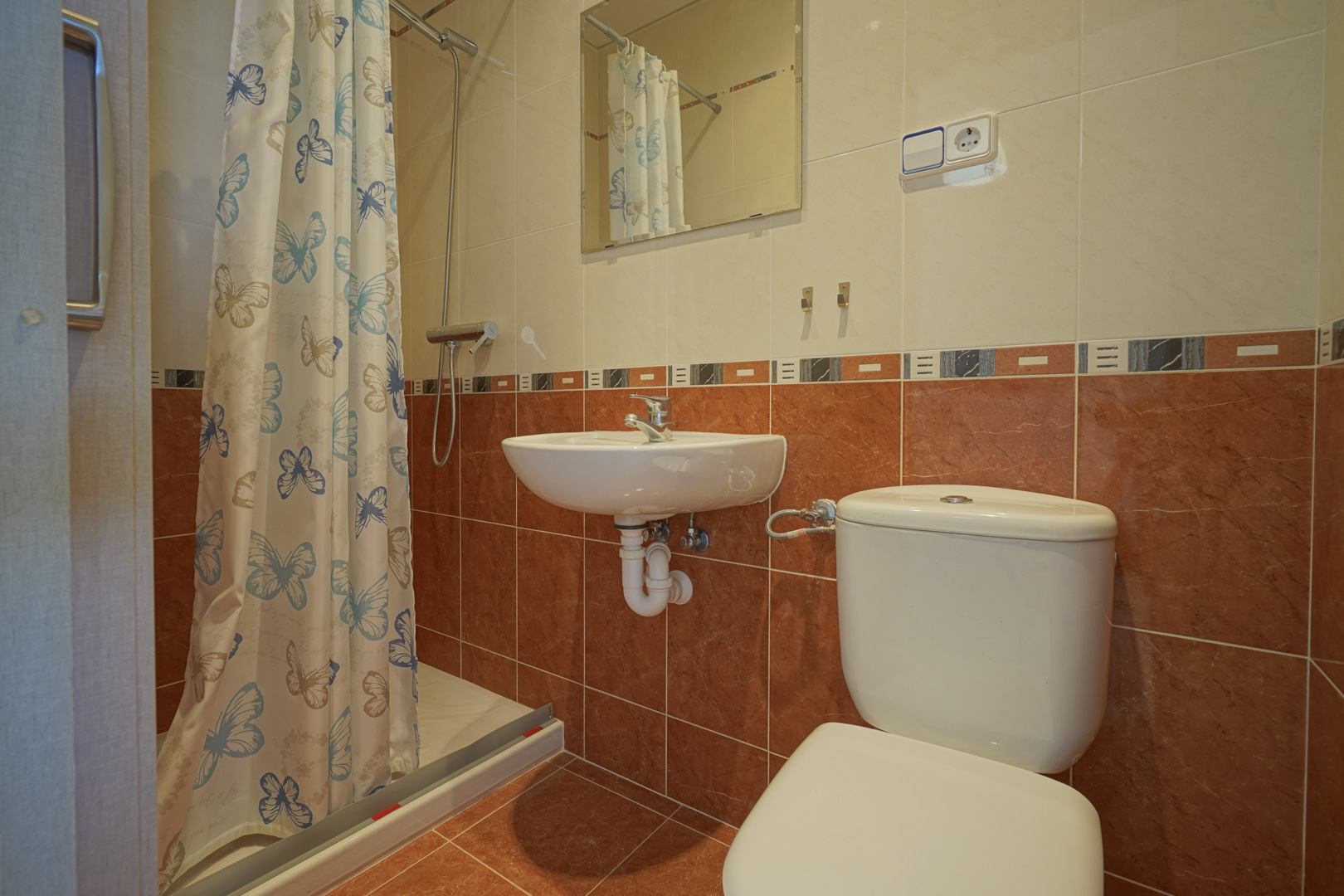 Cuarto de baño funcional con ducha, cortina de mariposas, lavabo de pared y azulejos en tonos terracota y crema con cenefa decorativa.