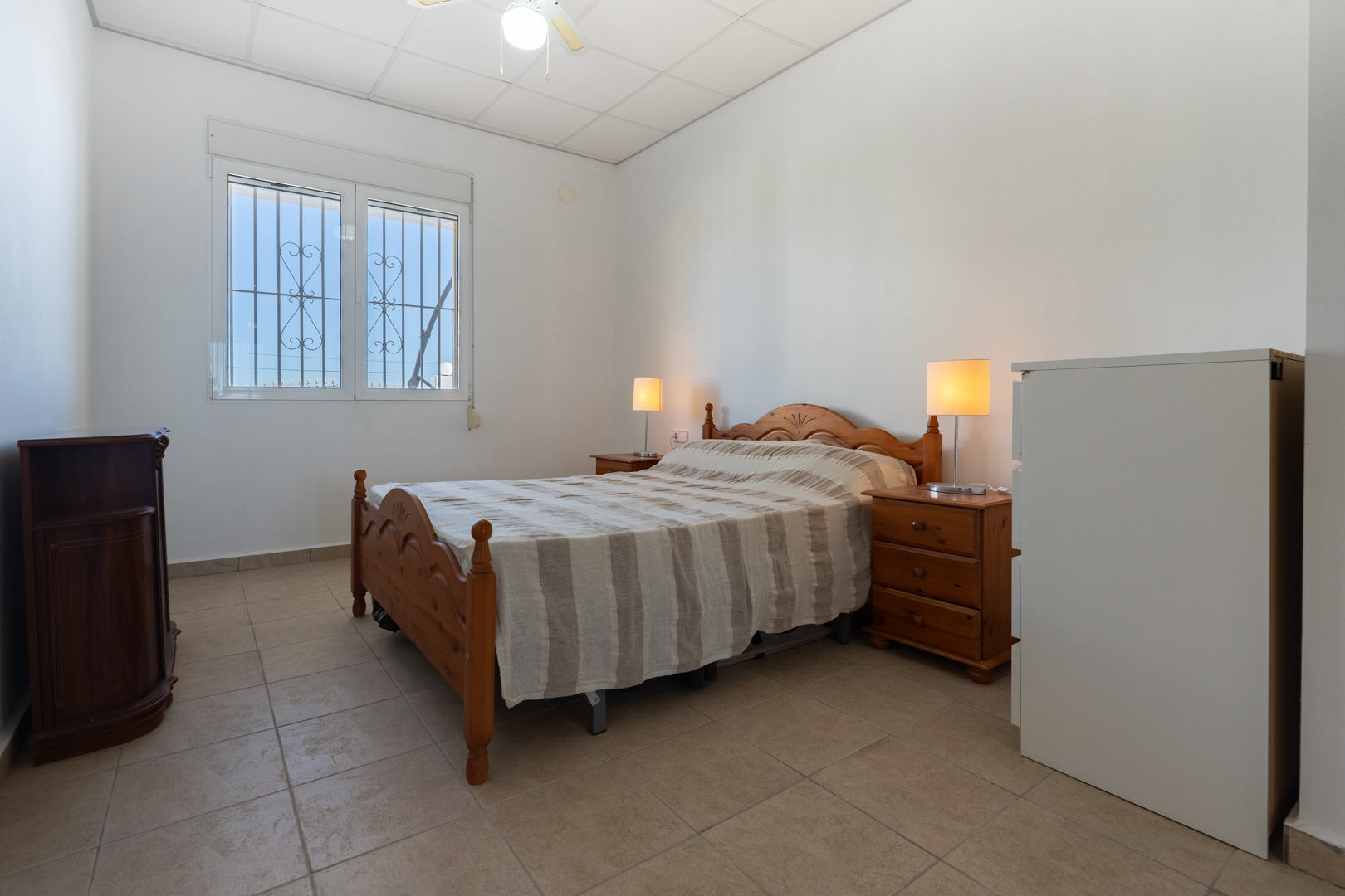 Dormitorio con mobiliario de madera tradicional, suelos de gres y ventana con persiana y rejas de seguridad que aporta luz natural.