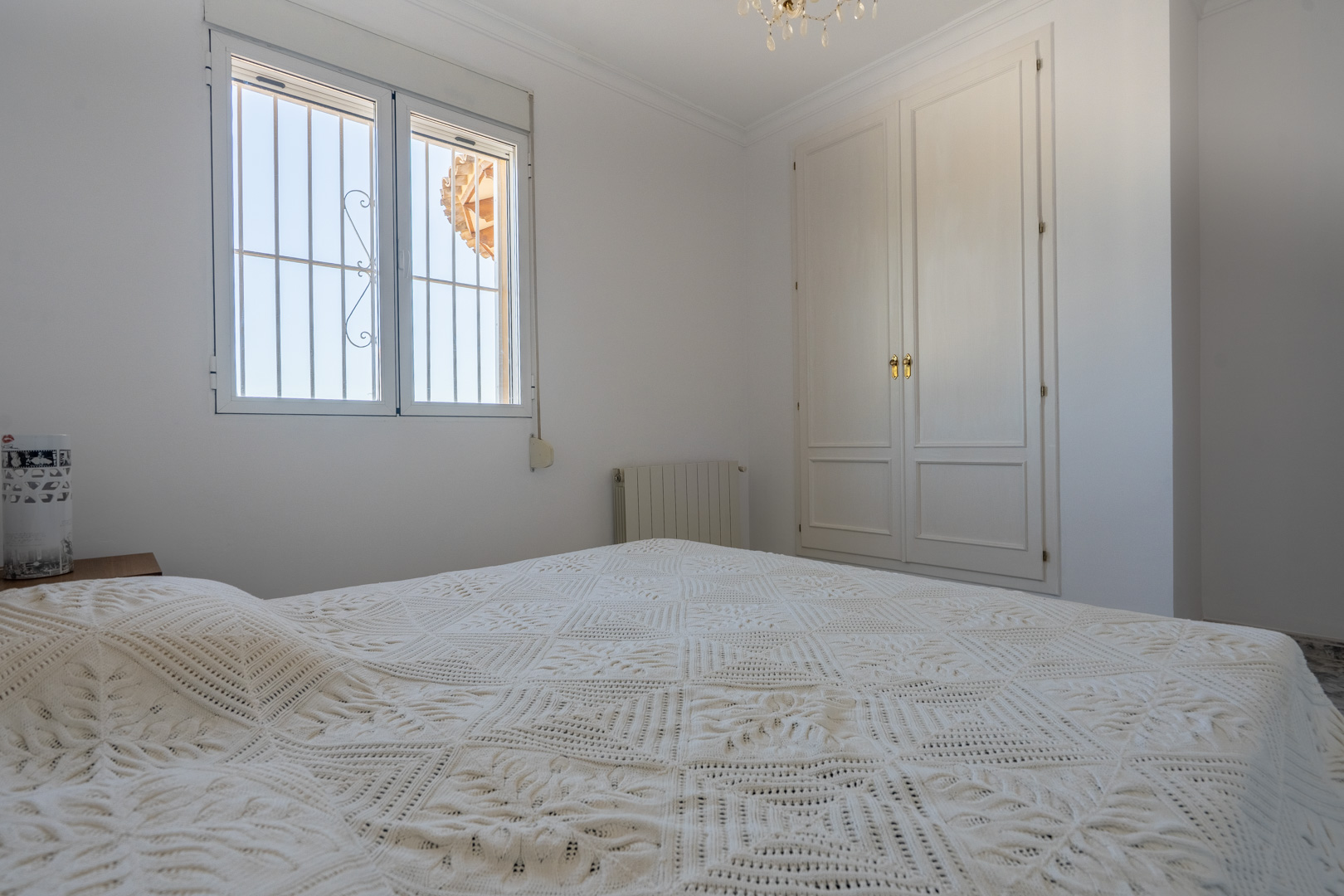Dormitorio luminoso con paredes blancas, armario empotrado de madera lacada y ventana con rejas de seguridad. Incluye calefacción por radiador y luz natural constante.