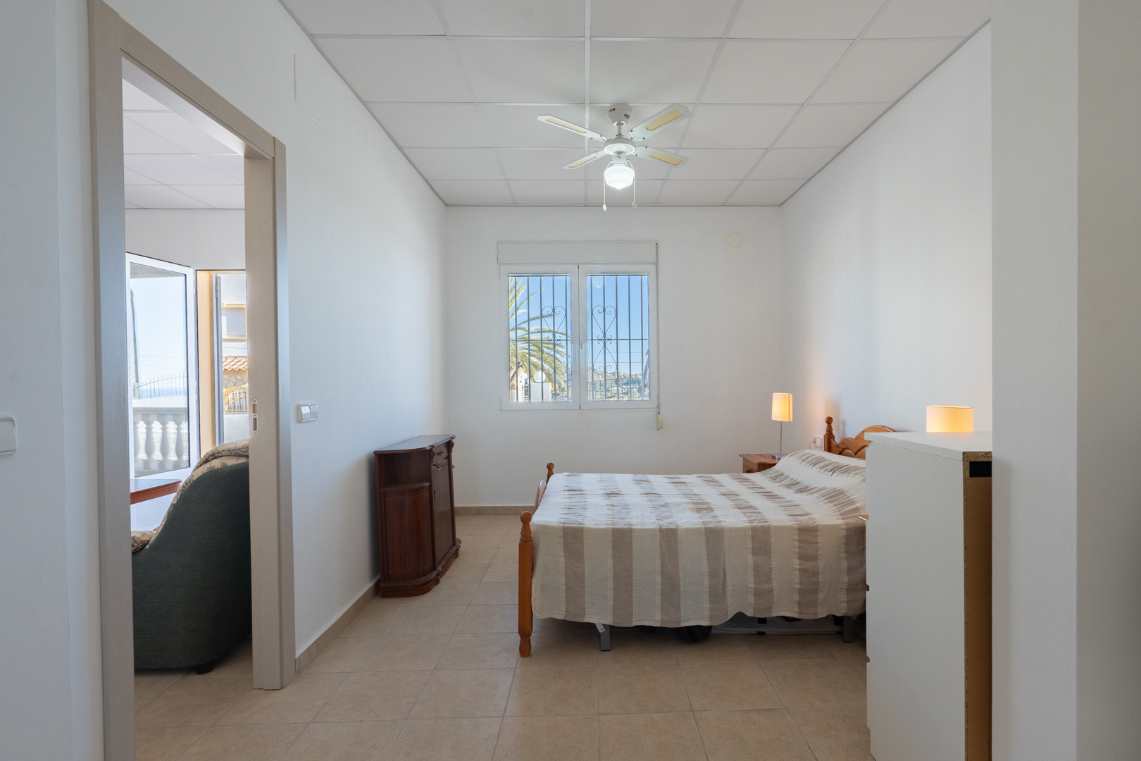 Dormitorio luminoso con suelos cerámicos, ventilador de techo y ventana con rejas. La estancia conecta con el salón y ofrece vistas a la vegetación exterior a través del ventanal.