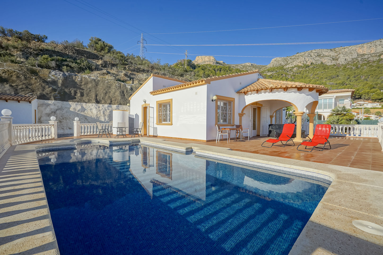 Exterior de la villa con piscina privada, amplia terraza de gres y porche cubierto con arcos, rodeado por el paisaje montañoso de la Sierra de Oltà.