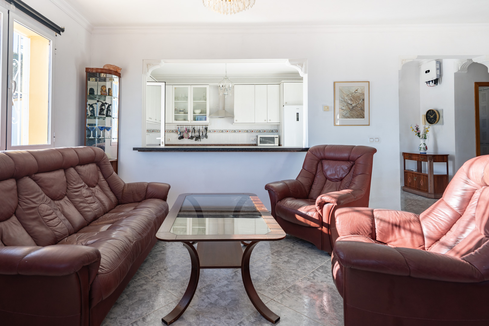 Salón con sofás de cuero marrón y suelos de gres efecto mármol, con una apertura decorativa que comunica visualmente con la cocina blanca equipada.