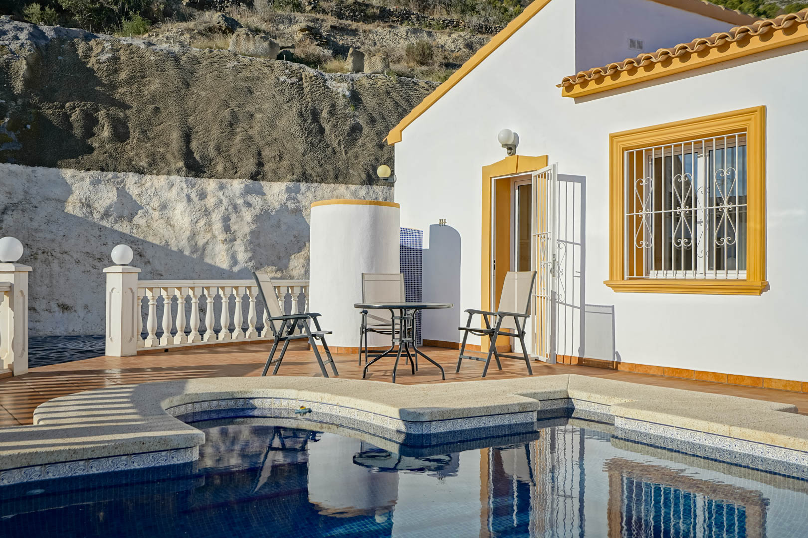 Terraza junto a la piscina con mobiliario exterior, ducha cilíndrica y fachada blanca con marcos ocres, situada frente a una ladera natural de piedra.