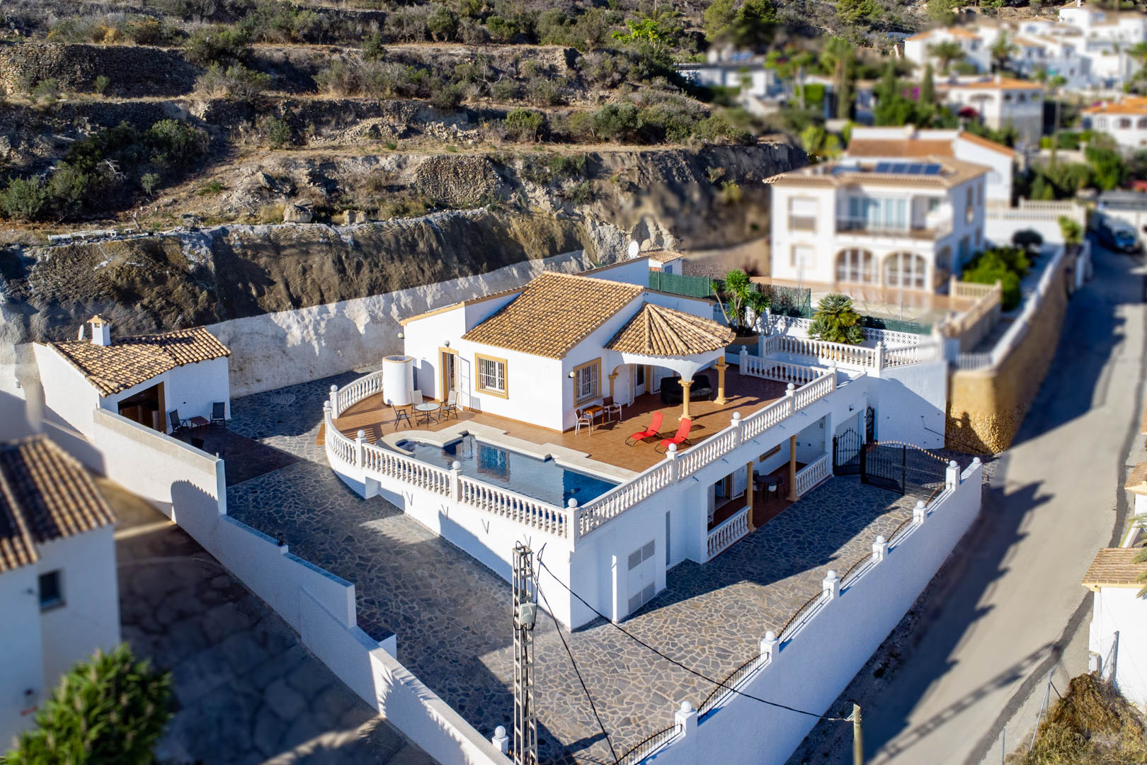 Vista aérea de la villa con piscina privada en terraza elevada, balaustrada blanca, tejados de teja y amplio patio pavimentado en piedra frente a la montaña.
