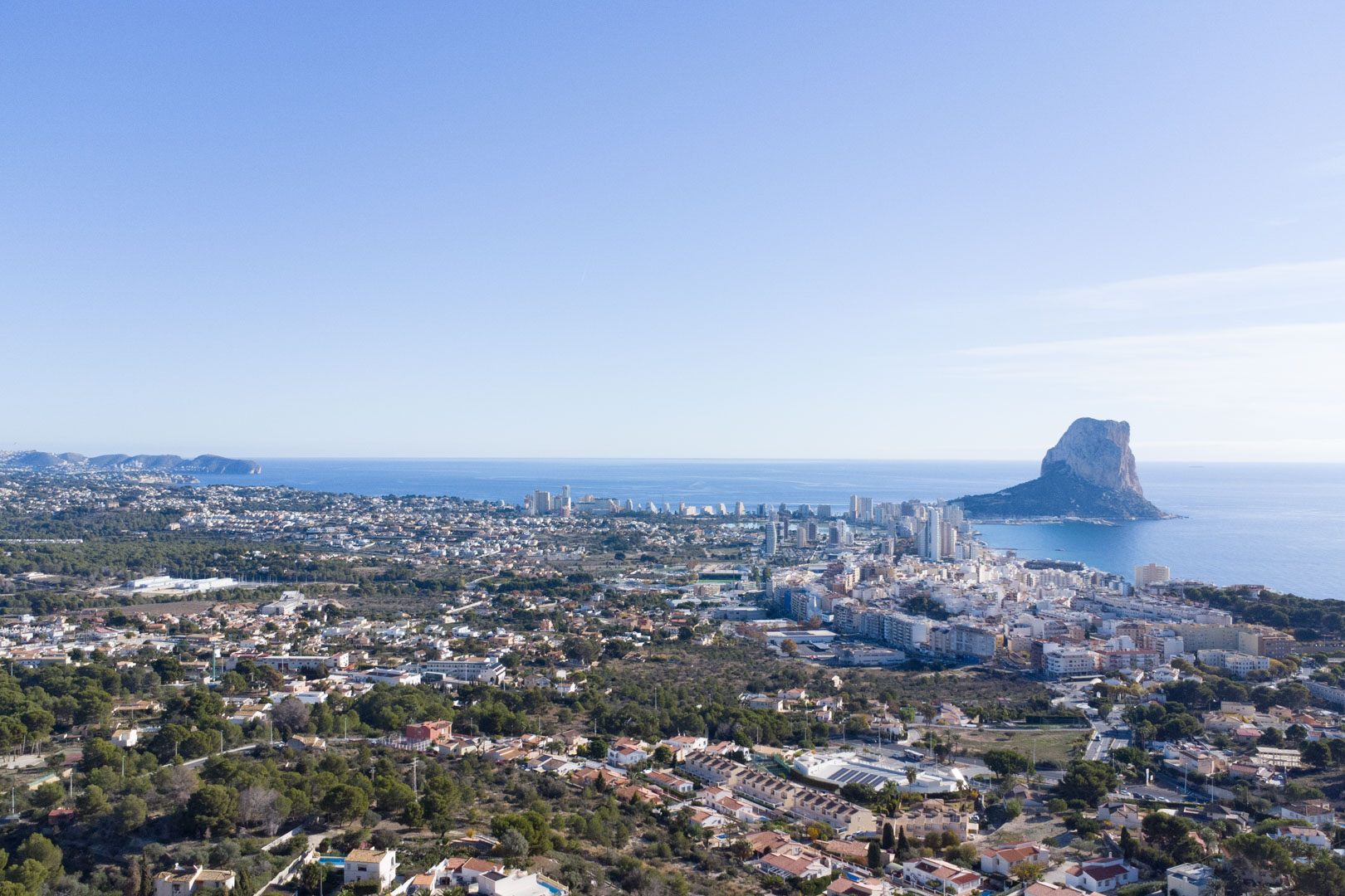 Vista aérea panorámica de la costa de Calpe, destacando el Peñón de Ifach, el skyline urbano y la extensión del mar Mediterráneo bajo un cielo despejado.