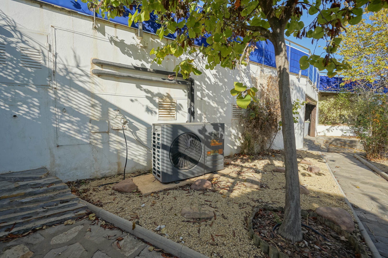 Área técnica exterior con bomba de calor Jetline Premium instalada sobre lecho de grava, junto a muros con instalaciones y zona ajardinada.