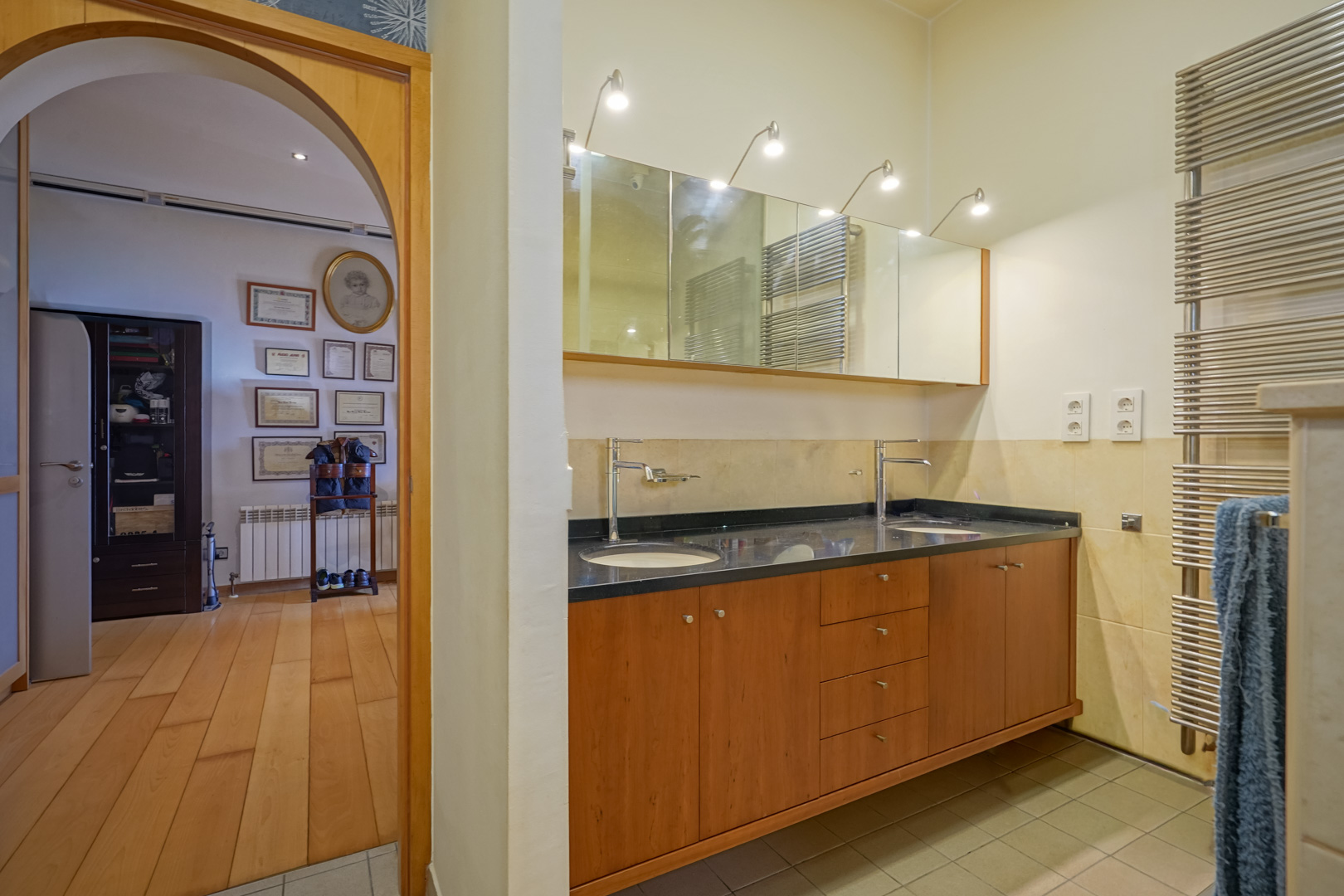 Baño con tocador doble de madera, encimera oscura y espejos iluminados. Se aprecia el acceso mediante un arco a un vestidor con suelos de madera noble.