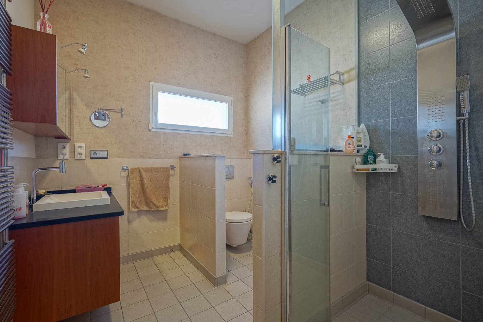 Baño funcional con columna de hidromasaje, mampara de cristal y zona de sanitarios separada. Incluye mueble de madera, encimera oscura y papel pintado con motivos de mapas.