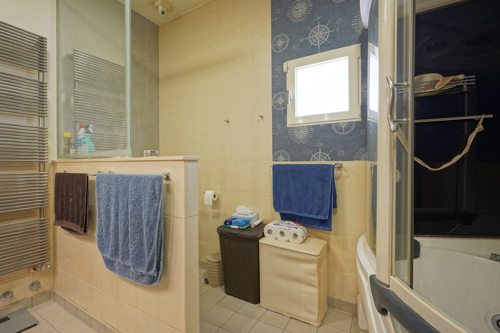 Baño funcional con revestimiento cerámico en tonos crema, radiador toallero de acero y zona de aseo independiente. Destaca el detalle decorativo náutico junto a la ventana.