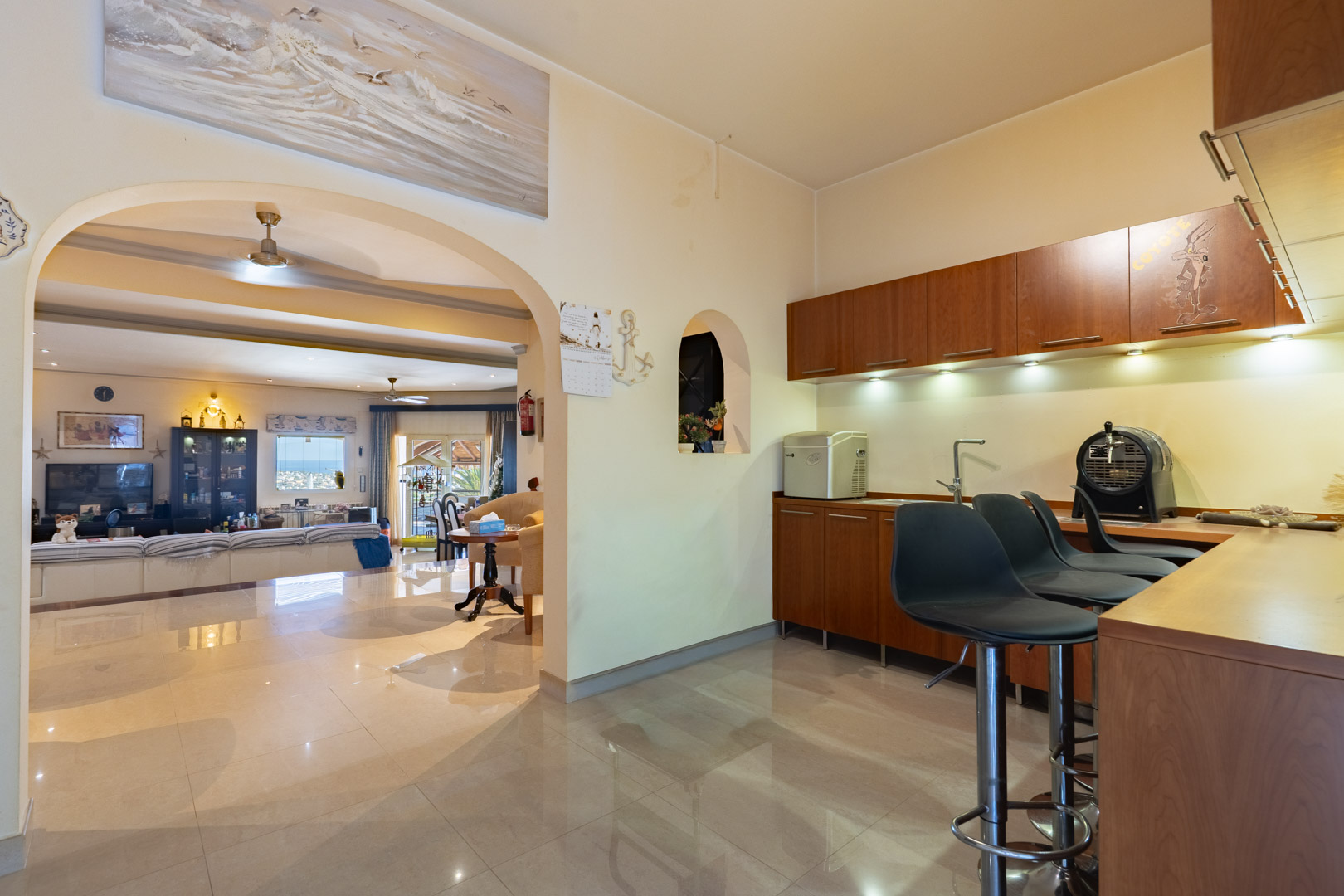 Bar integrado con mobiliario de madera, taburetes altos e iluminación bajo los armarios, conectado al salón principal mediante un arco y suelos cerámicos de brillo.