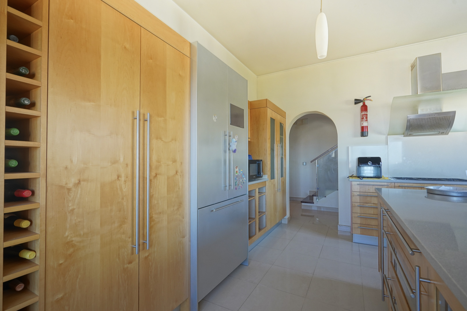 Cocina con mobiliario de madera a medida, botellero integrado y electrodomésticos de acero inoxidable. El arco comunica con la zona de escaleras con barandilla de cristal.