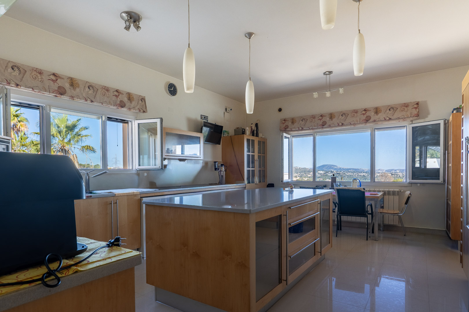Cocina espaciosa con isla central, mobiliario de madera y encimeras grises. Grandes ventanales ofrecen vistas despejadas al mar y la costa de Calpe bajo luz natural.
