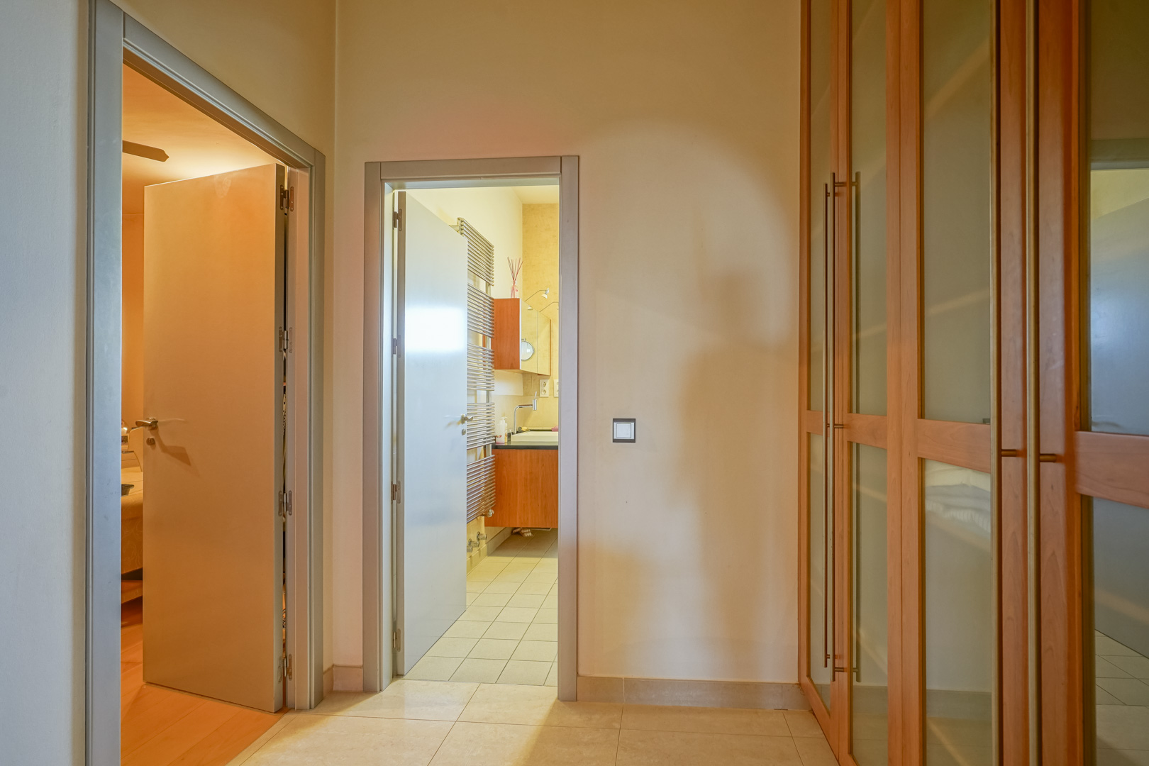 Distribuidor funcional con armarios empotrados de madera y cristal, que conecta un dormitorio de suelos nobles con un baño en suite equipado con radiador toallero.