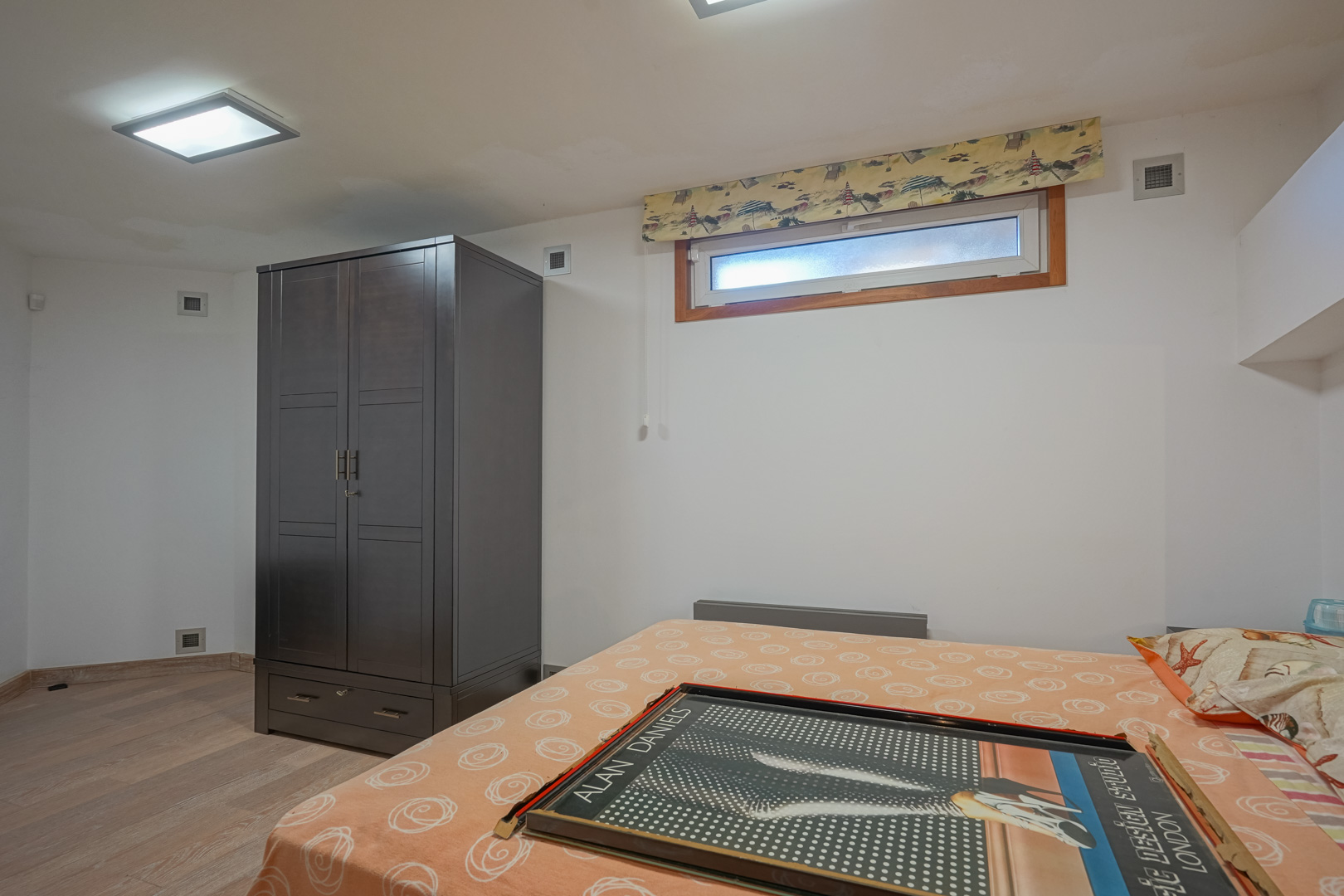 Dormitorio funcional con armario de madera oscura, suelos laminados claros y una ventana horizontal que aporta luz natural bajo el techo con focos empotrados.
