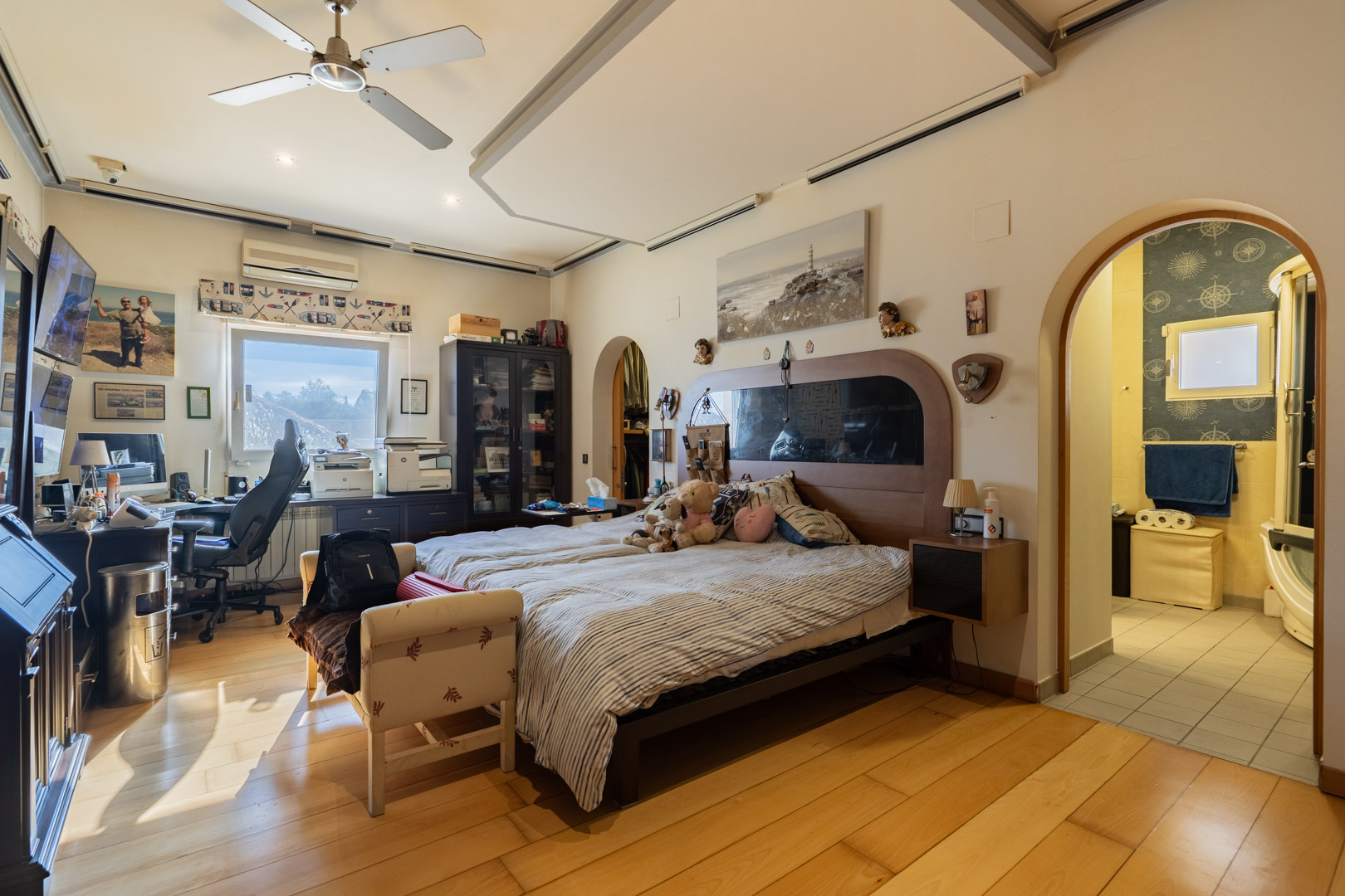 Dormitorio principal con suelos de madera noble, zona de despacho integrada y baño en suite accesible mediante un arco. Incluye ventilador de techo y aire acondicionado.