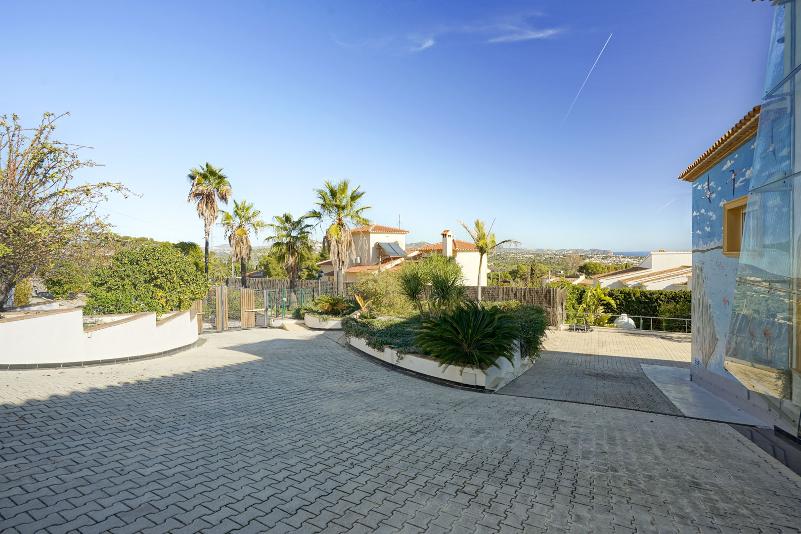 Entrada principal pavimentada con adoquines, rodeada de zonas ajardinadas con palmeras y un mural artístico lateral, ofreciendo vistas despejadas hacia la costa de Calpe.