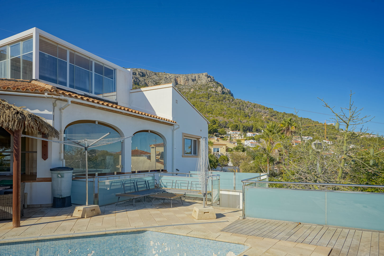 Exterior de la villa con terraza de gres, barandillas de cristal y vistas a la Sierra de Oltamar. Destacan los ventanales arqueados y el cerramiento acristalado superior.