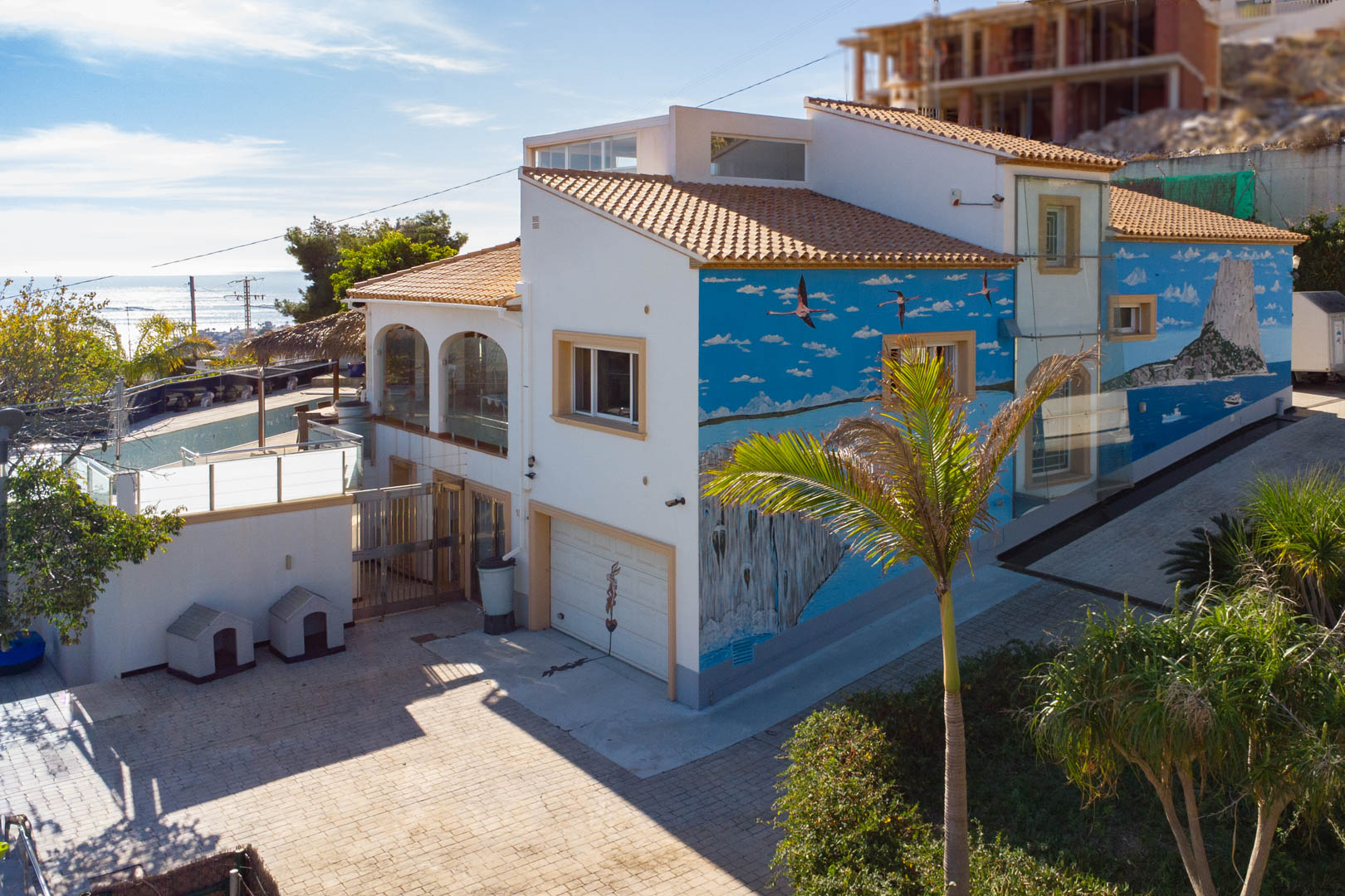 Exterior de la villa con un mural artístico del Peñón de Ifach, acceso al garaje y terrazas con cerramientos de cristal bajo un tejado de teja tradicional.
