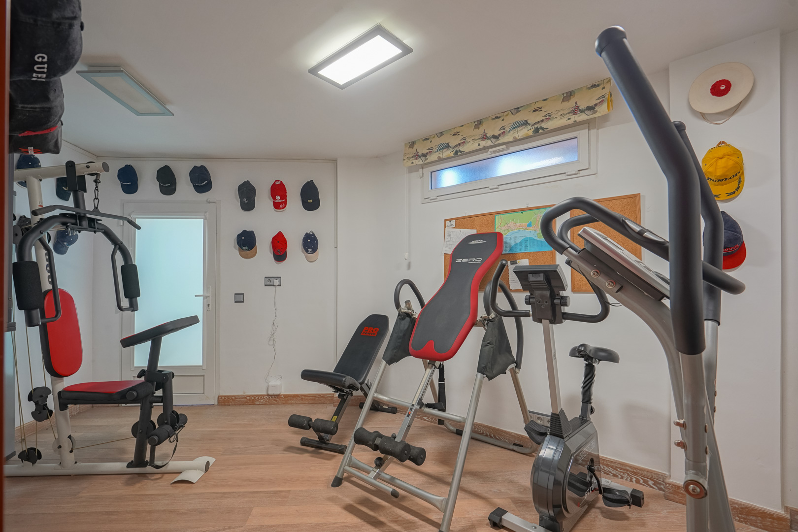 Gimnasio privado con suelo de madera, equipado con máquina multiestación, bicicleta estática y tabla de inversión bajo iluminación de paneles LED.