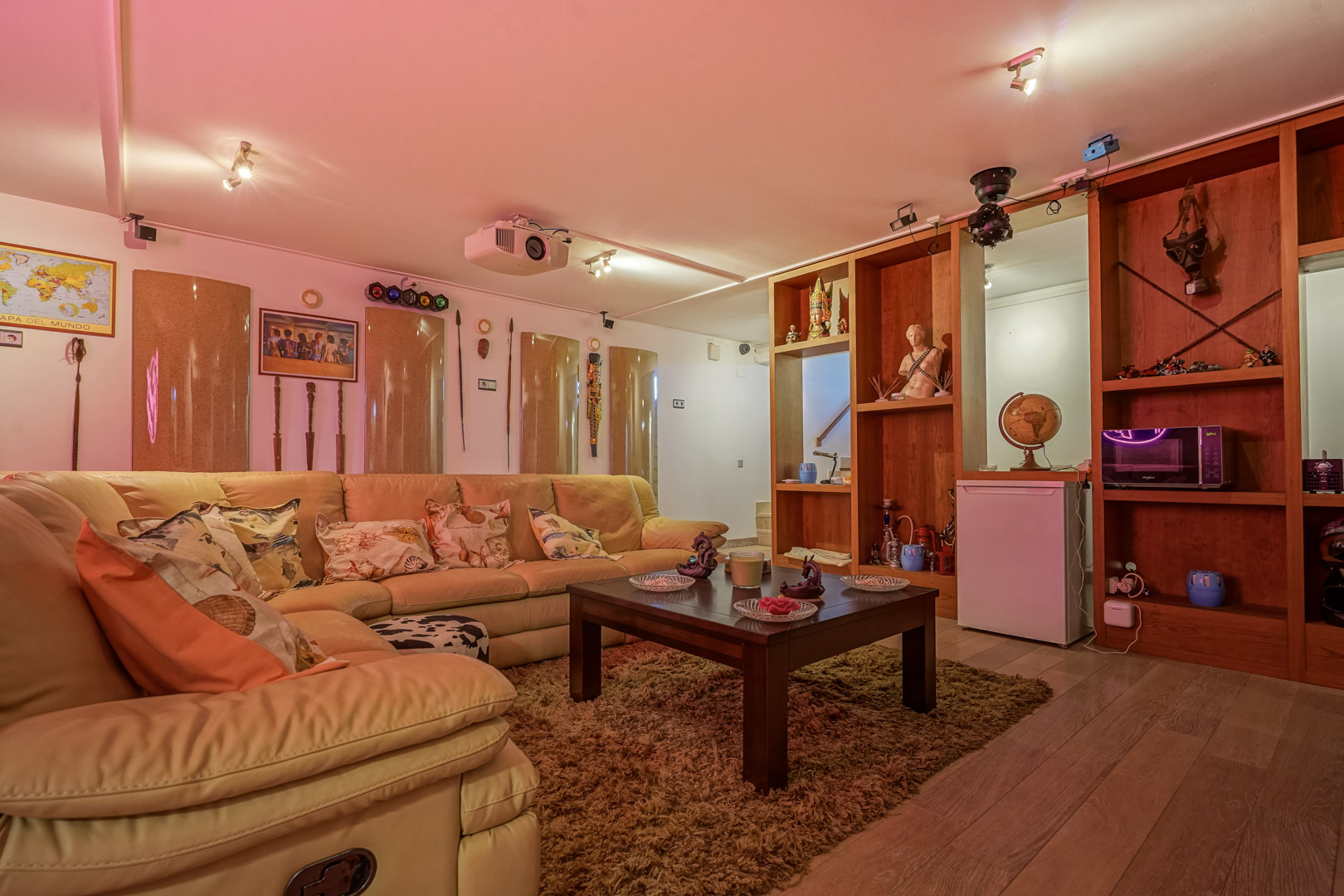 Sala multimedia con sofá rinconero de cuero, proyector de techo, luces de discoteca y estantería de madera integrada con zona de bar y pequeños electrodomésticos.