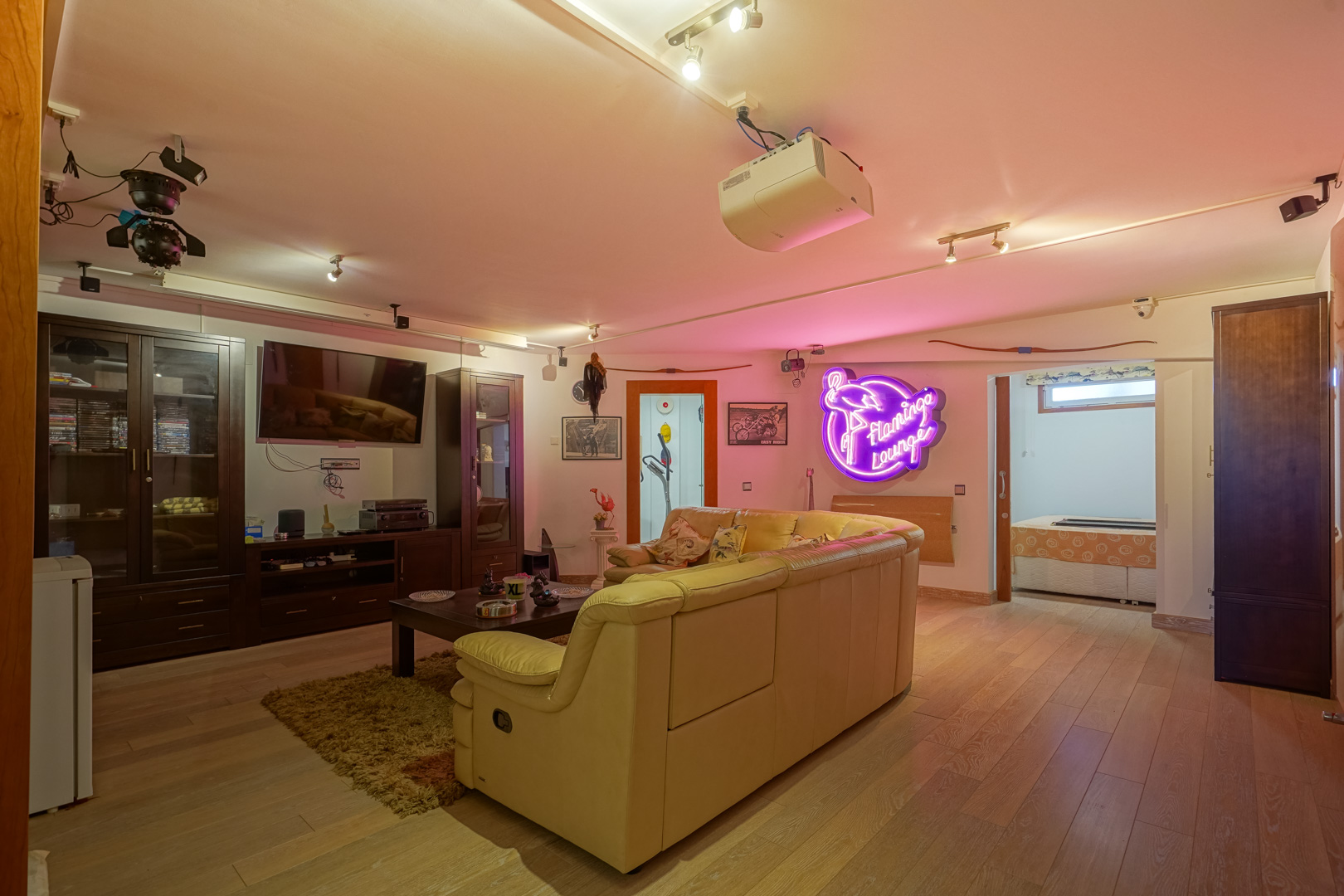 Sala multimedia con suelos de madera, proyector de techo y zona de estar con sofá de cuero. Incluye mobiliario oscuro, iluminación técnica y un distintivo letrero de neón.