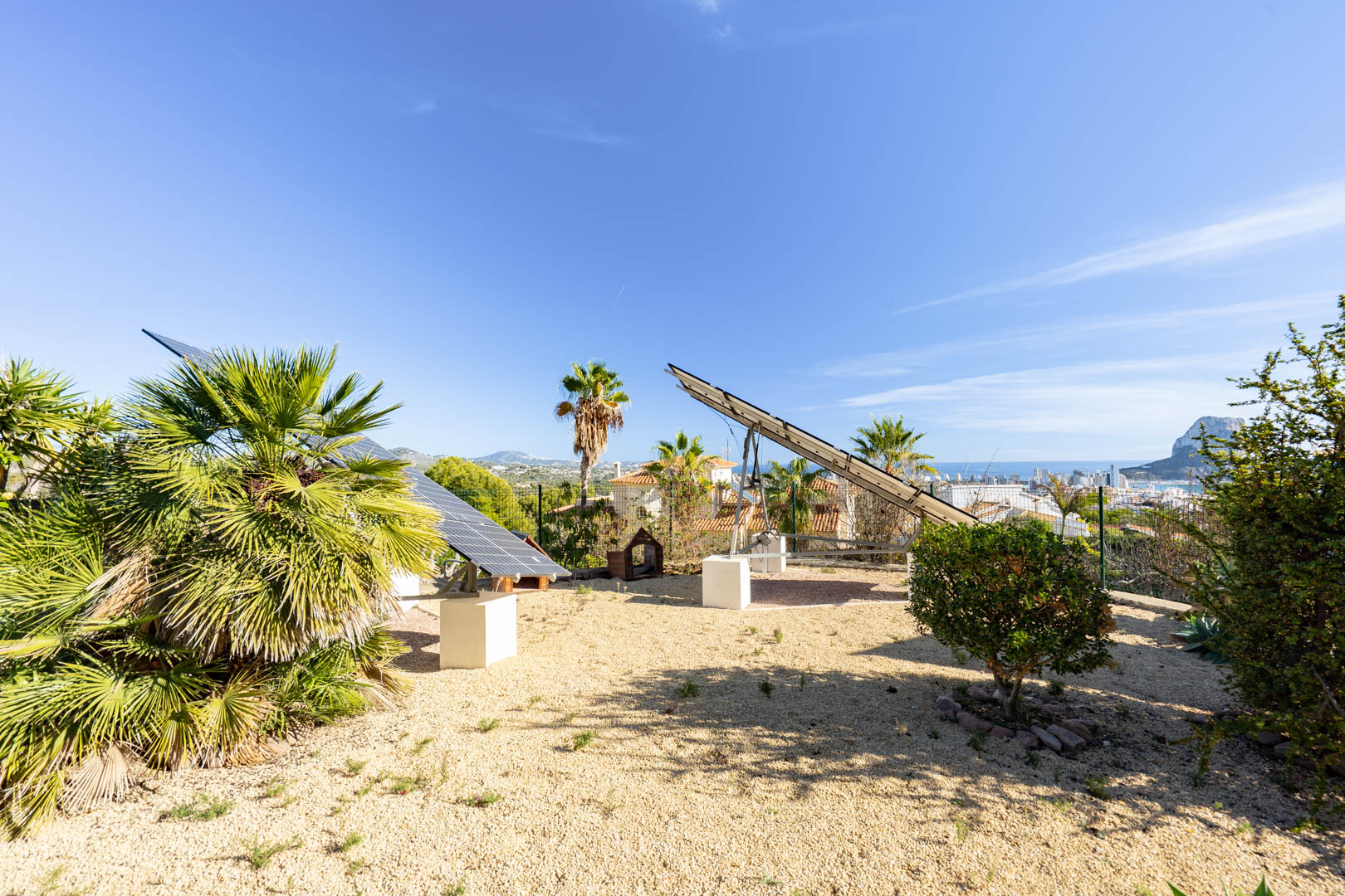 Sistema de placas solares motorizadas sobre bases de hormigón en zona de grava, con vistas panorámicas al Peñón de Ifach y el skyline de Calpe.