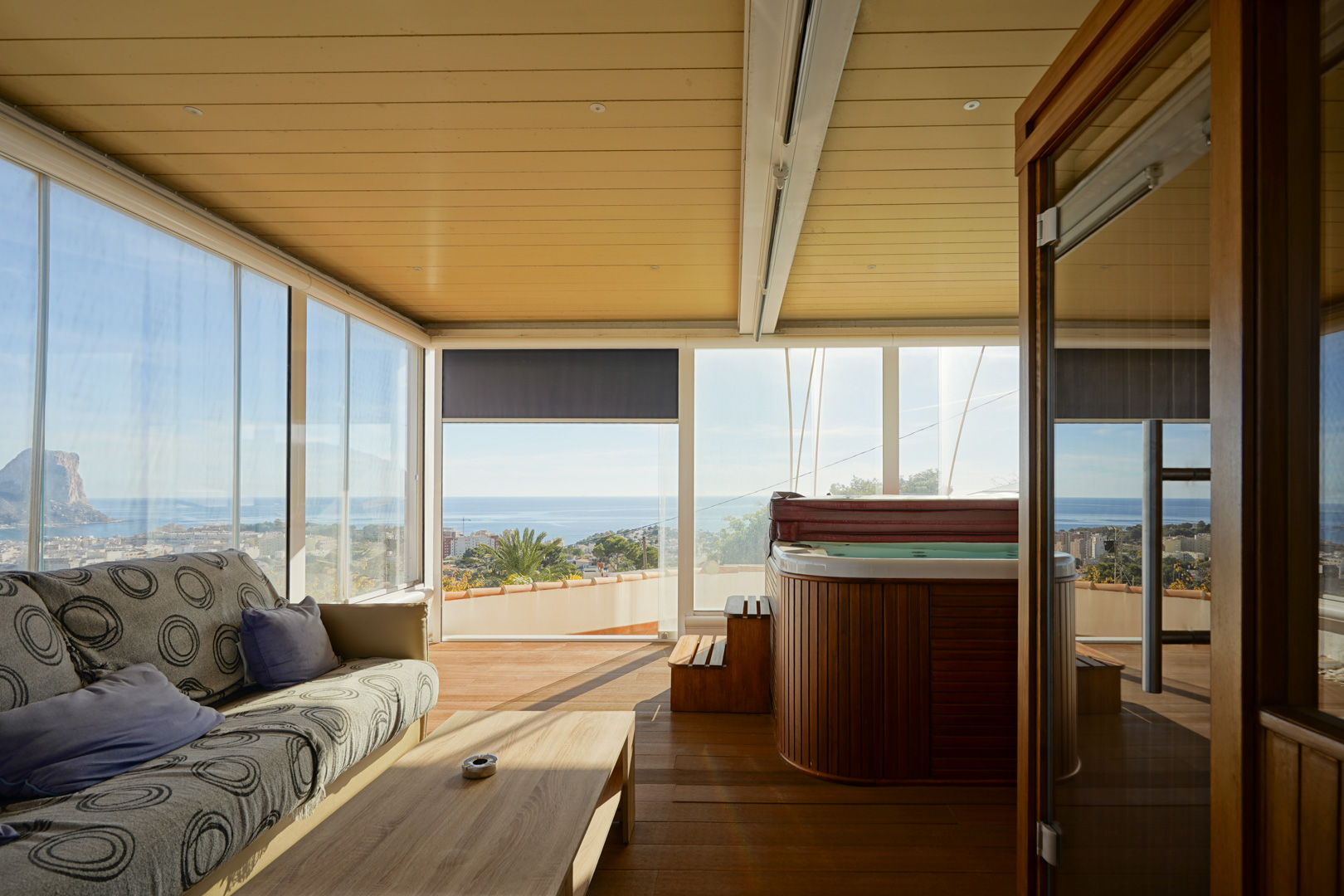 Terraza acristalada con jacuzzi revestido en madera, zona de estar y vistas panorámicas al Peñón de Ifach. Cuenta con suelos de madera y techo con iluminación integrada.